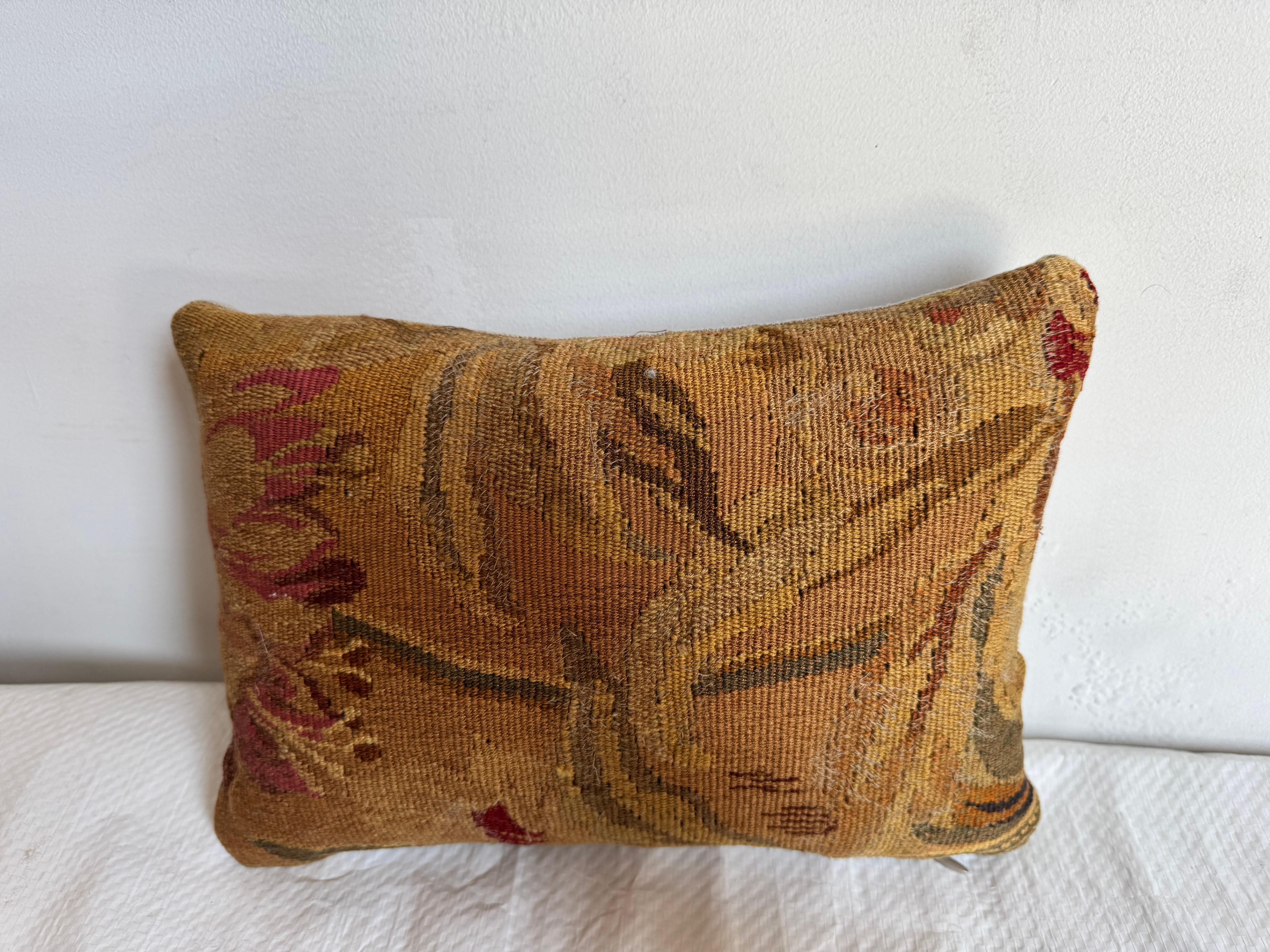 Se trata de un pequeño cojín tapiz vintage de estilo Aubusson con un motivo floral o botánico abstracto. El diseño está realizado en una cálida paleta otoñal de ocres, dorados apagados, olivos y burdeos desvaído, tejida en una superficie plana y