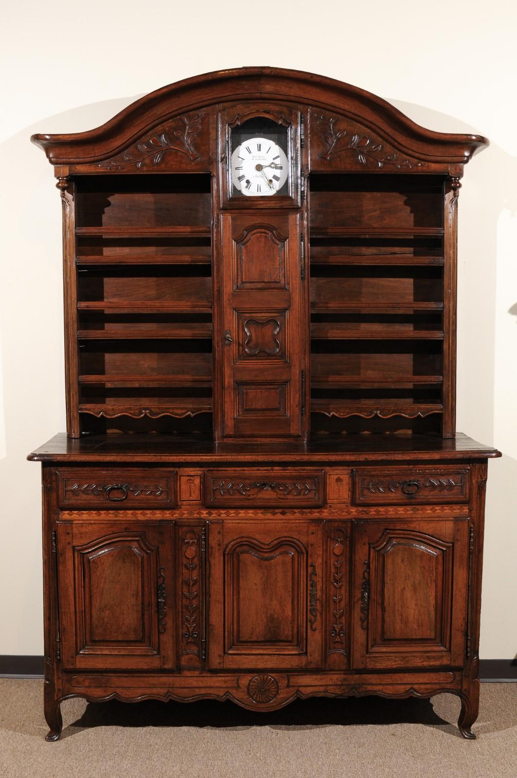 A French 18th century vaisellier in walnut featuring arched cornice, plate rack with enclosed clock workings, 3 paneled door cabinet below with foliage carving and parquetry inlay and shaped apron ending on cabriole feet. The Vaisellier has