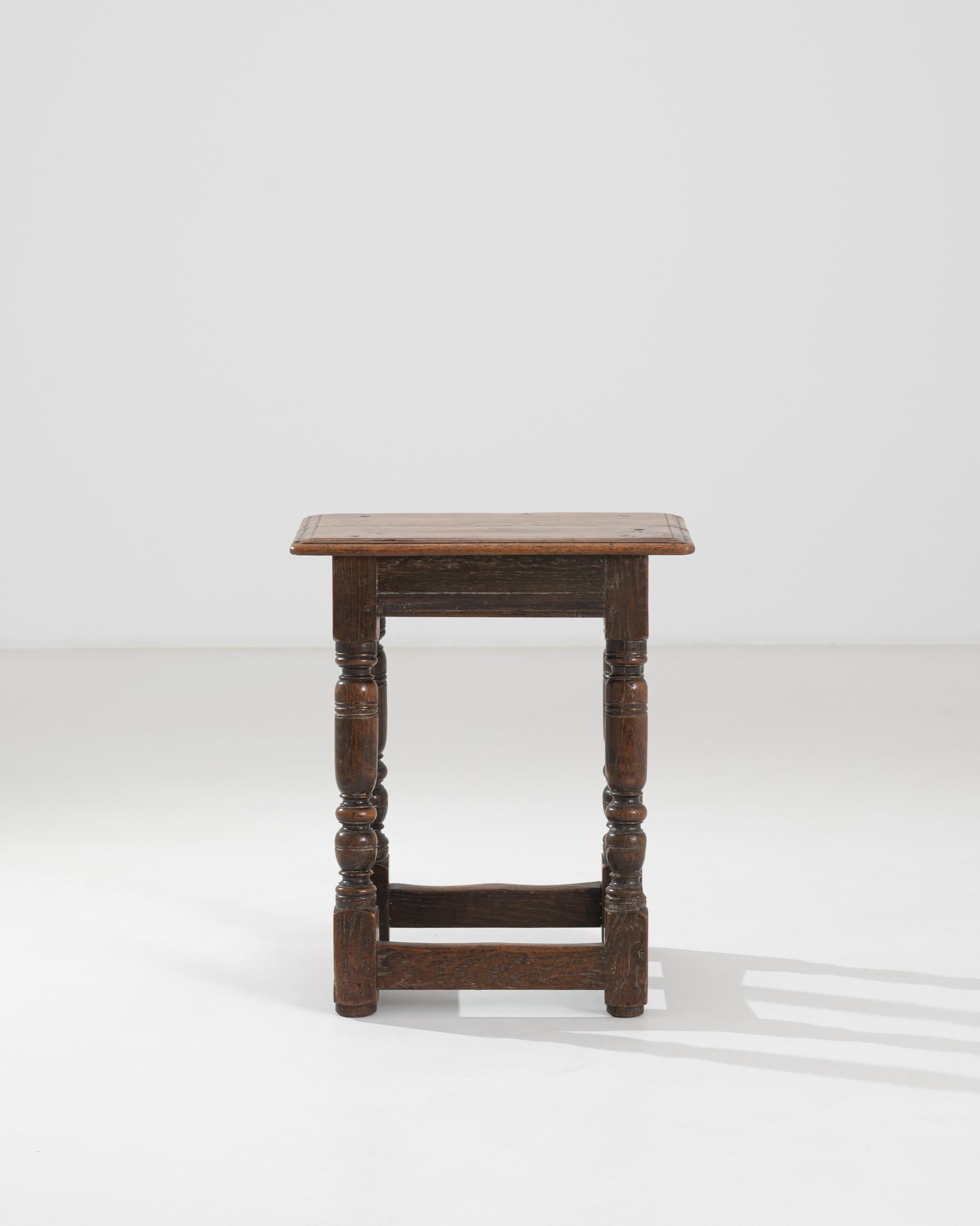 An oak stool produced in France. The piece features angled legs, joined by square stretchers and assembled with mortise and tenon joints. The wood is colored with its original finish, the natural tone of the oak, highlighting a discrete dark patina.