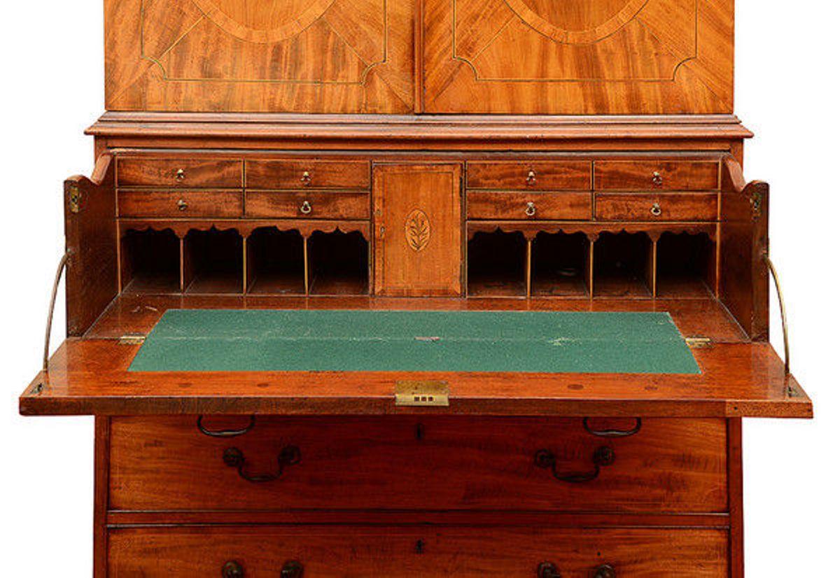 A Geo 111 mahogany and satinwood inlaid secretaire linen press.
The top with a pierced swan-neck pediment and dentil cornice and a pair of crossbanded panel doors which open to reveal three sliding trays.
The secretaire with a false front of two