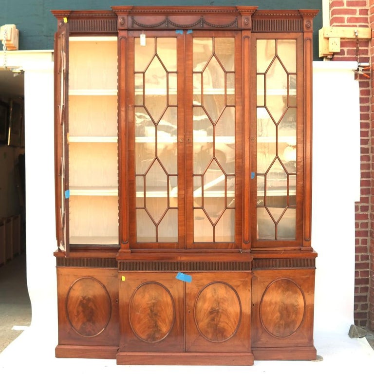 George III mahogany breakfront bookcase, the pierced, scrolled broken pediment above a partially inlaid, stepped molding, above a fluted frieze centered by foliate swags, with paterae to the four corners, above a pair of glass mullioned cabinet