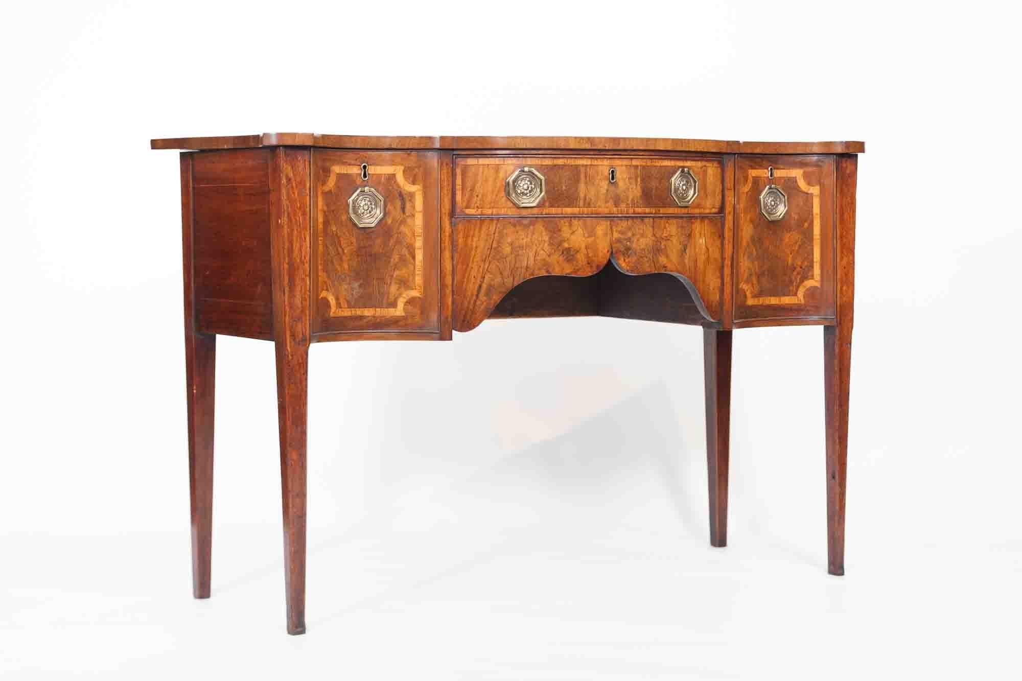 18th century George III walnut serpentine sideboard attributed to Thomas Sheraton, the shaped moulded top raised over single cross banded cockbeaded short drawer with satinwood inlay and hexagonal brass pulls with rosehead motif and brass escutcheon