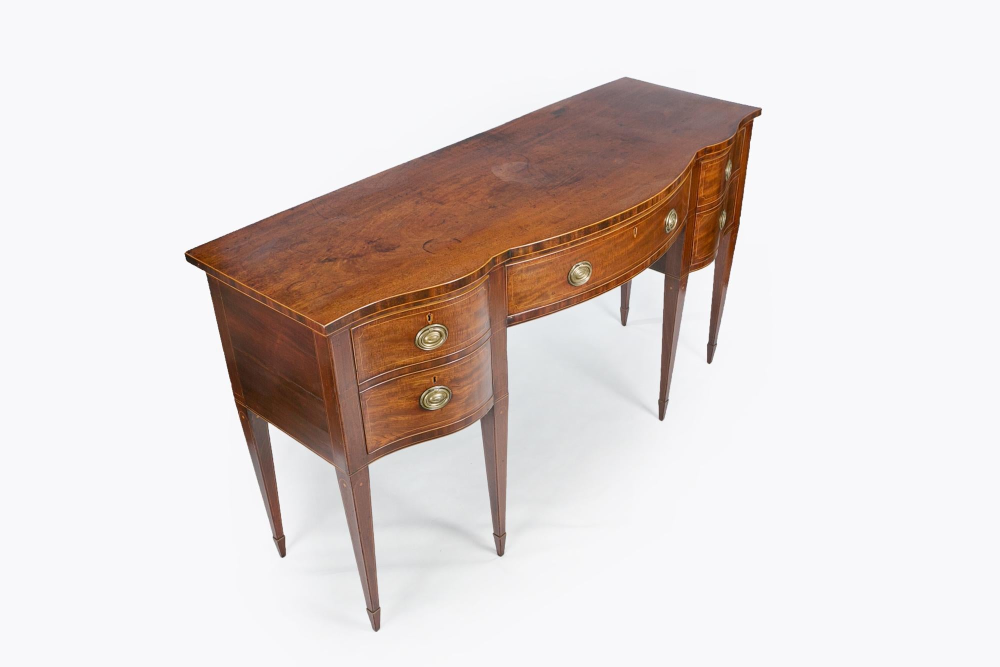 18th century George III mahogany serpentine fronted sideboard, the moulded top with satinwood line inlay raised over central bow shaped cockbeaded drawer frieze flanked with pair of cellarete configured cockbeaded drawers, all with decorative brass