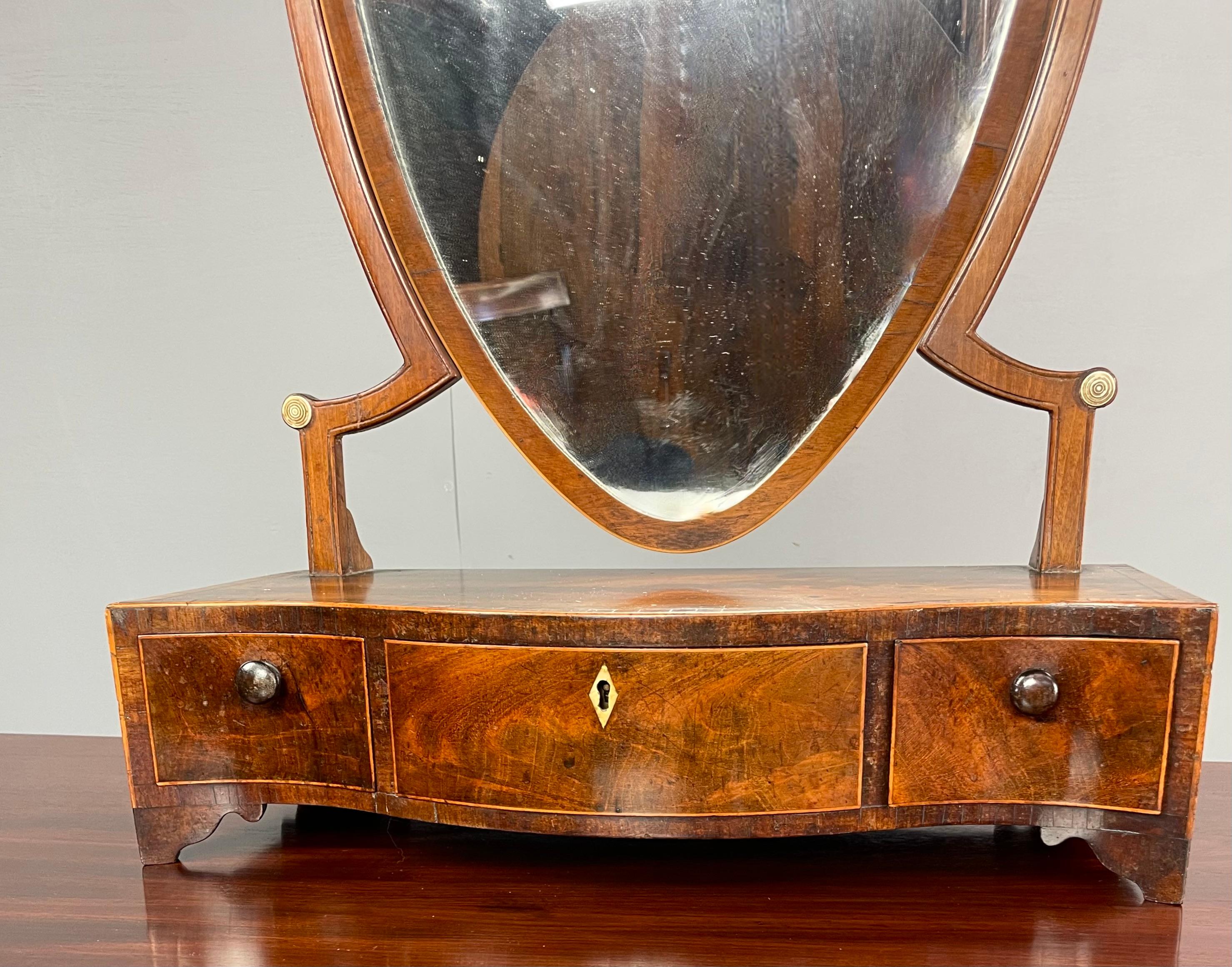 A Sheraton period, late 18th century, serpentine fronted, 3 drawer toilet mirror having a shield shaped glass.
This is a lovely proportioned little mirror with good figuring and boxwood stringing standing on ogee bracket feet.
The swing mirror is
