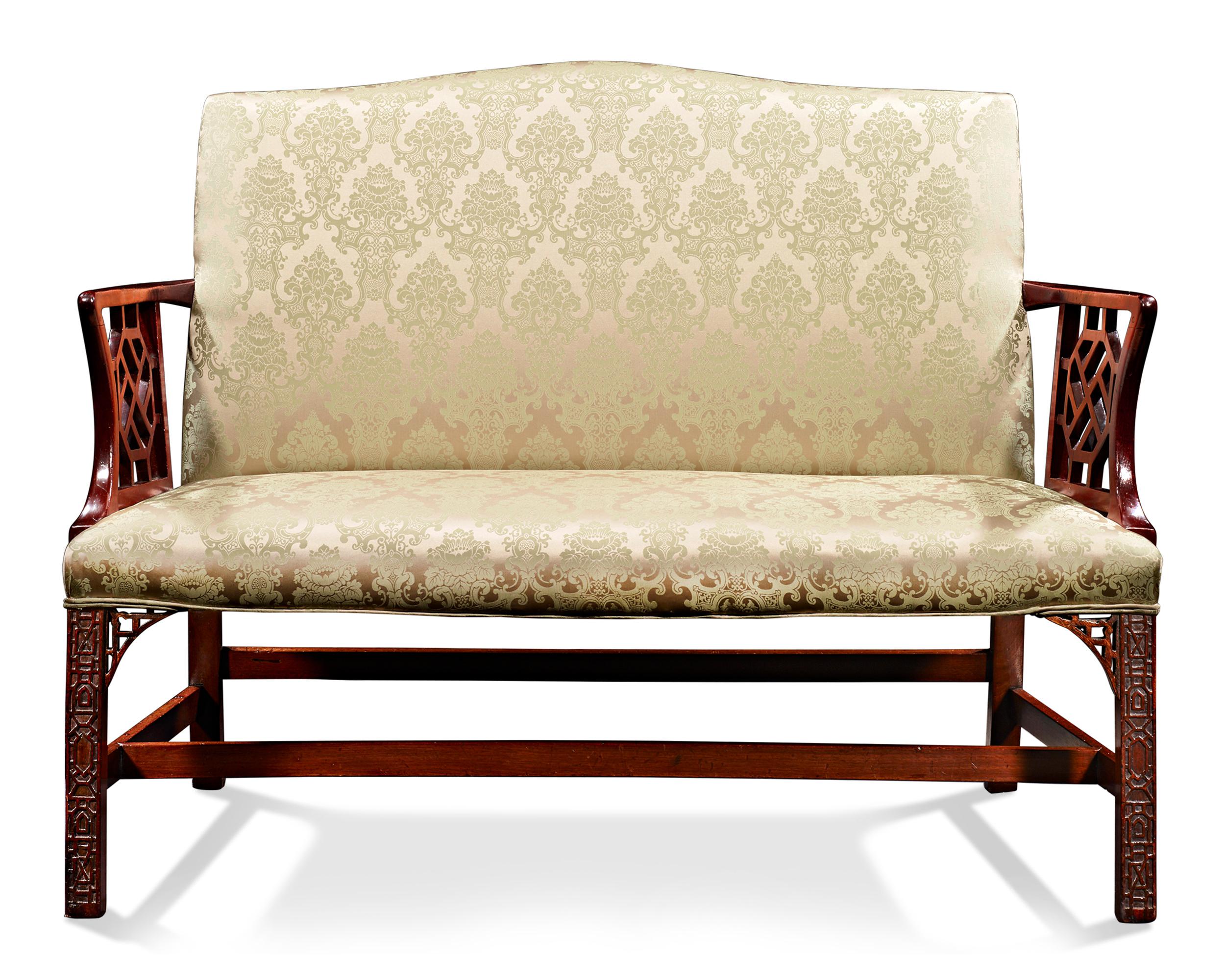 This beautiful George III-era upholstered settee features delicate serpentine lines highlighted by luxurious mahogany. Open trellis arms and square legs carved with Fine blind fretwork are headed by pierced brackets and joined by an H-formed