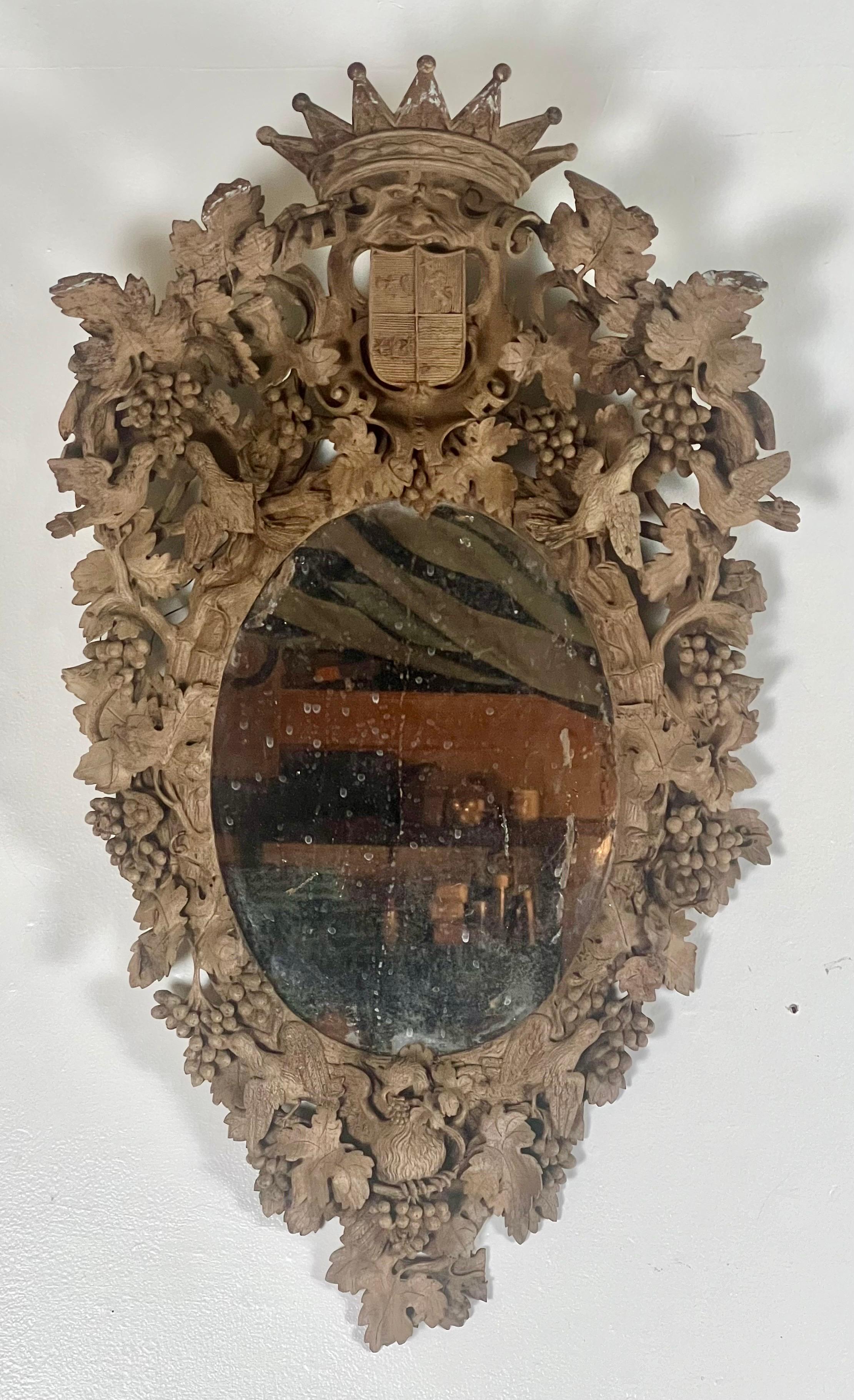 18th century German mirror, finely detailed with a crown, coat of arms, vines with grapes and leaves, and petite birds, all surrounding an oval-shaped mirror.