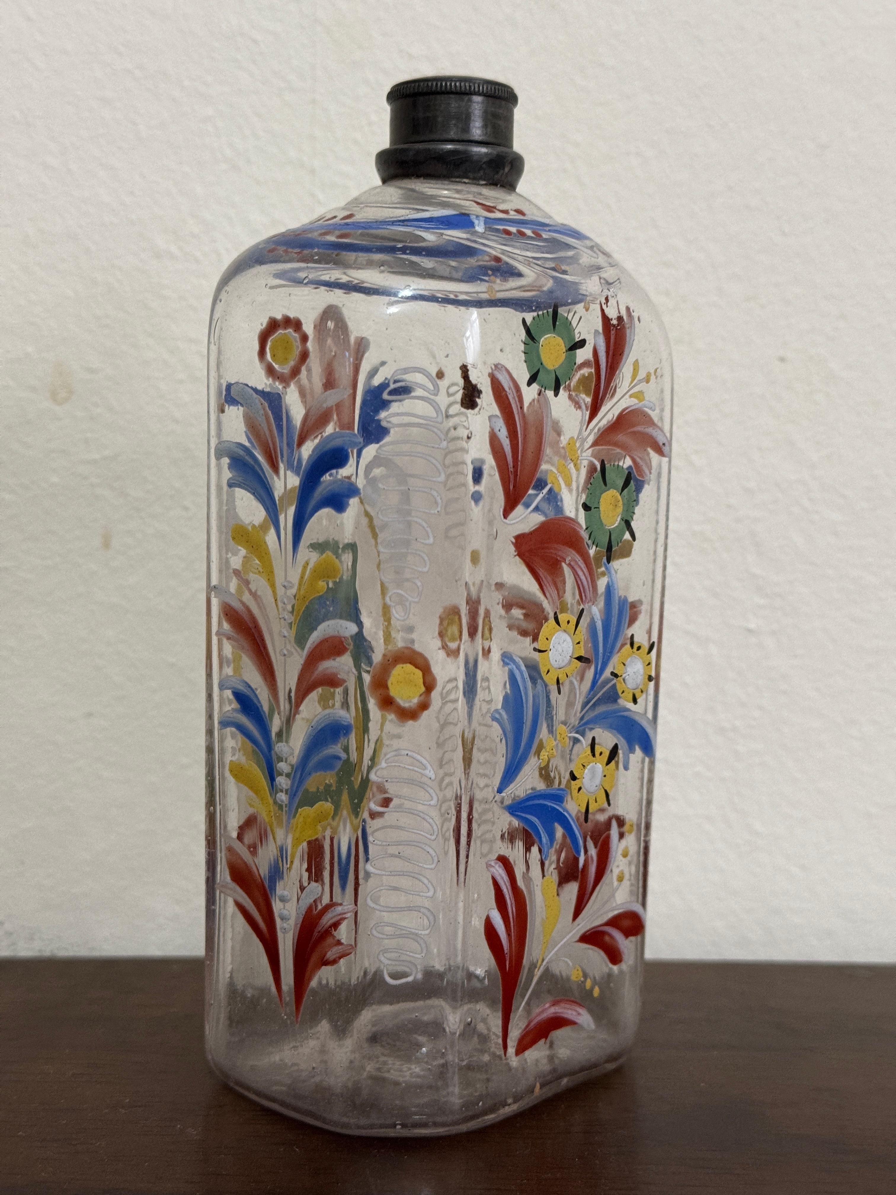 Bottiglia di vetro trasparente del XVIII secolo, tedesca o forse boema. Forma ottagonale appiattita. Decorata a mano con fiori blu, arancioni, gialli e verdi e un  uccello verde/blu sul retro.

15cm / 6