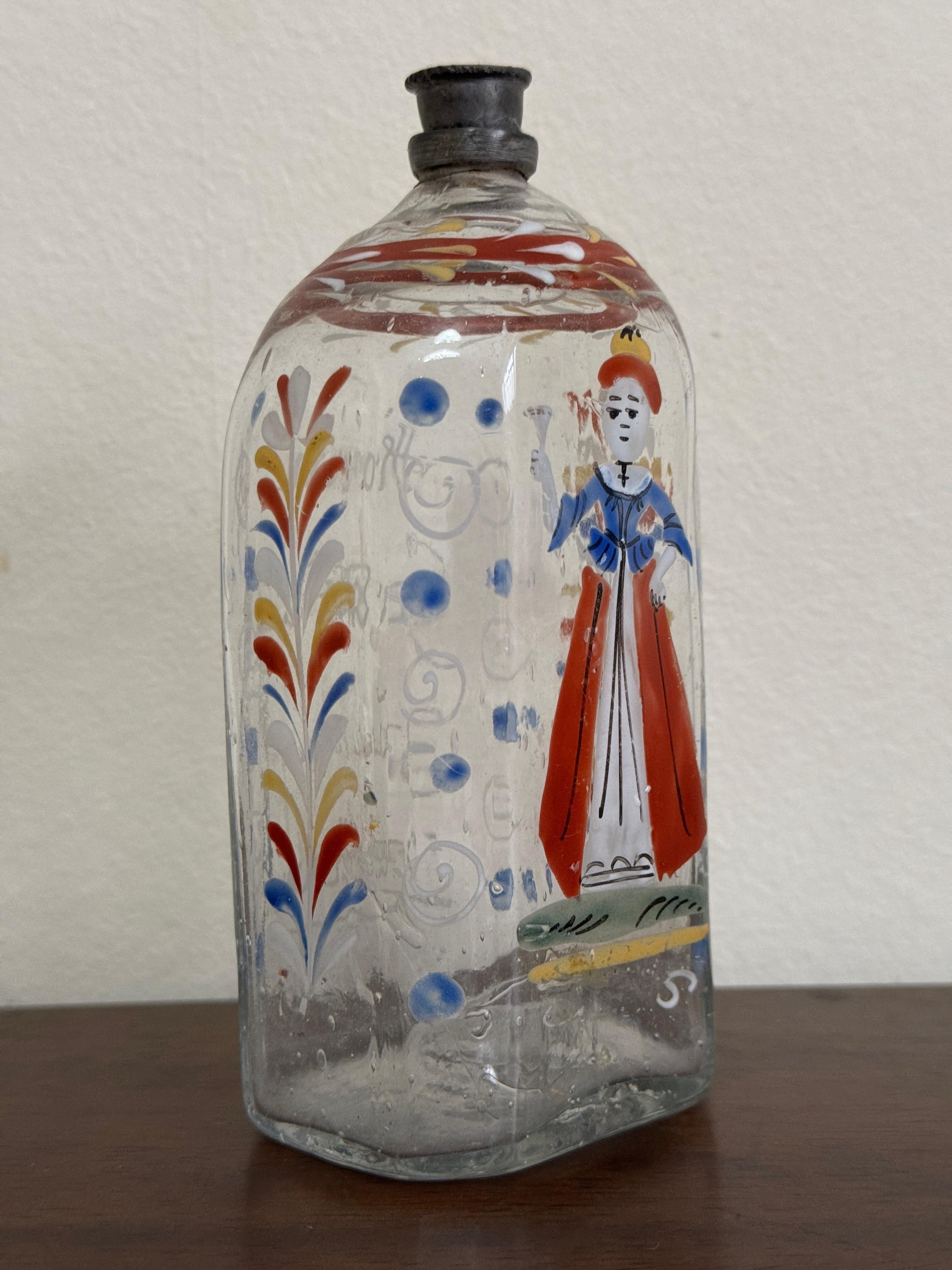 Bottiglia di vetro trasparente del XVIII secolo, tedesca o forse boema. Forma ottagonale appiattita. Decorata a mano con fiori blu, arancioni, gialli e bianchi e una ragazza con una camicetta blu e un vestito rosso sul davanti. Testo tedesco