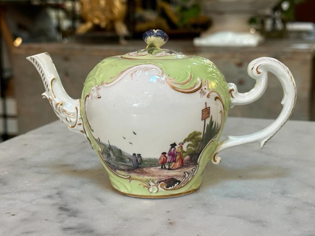 Meissen teapot decorated with countryside and riverside scenes populated by traveling figures and framed by raised and gilt foliage designs against a pastel green background embellished with floral motifs and raised foliage designs. Spout and handle