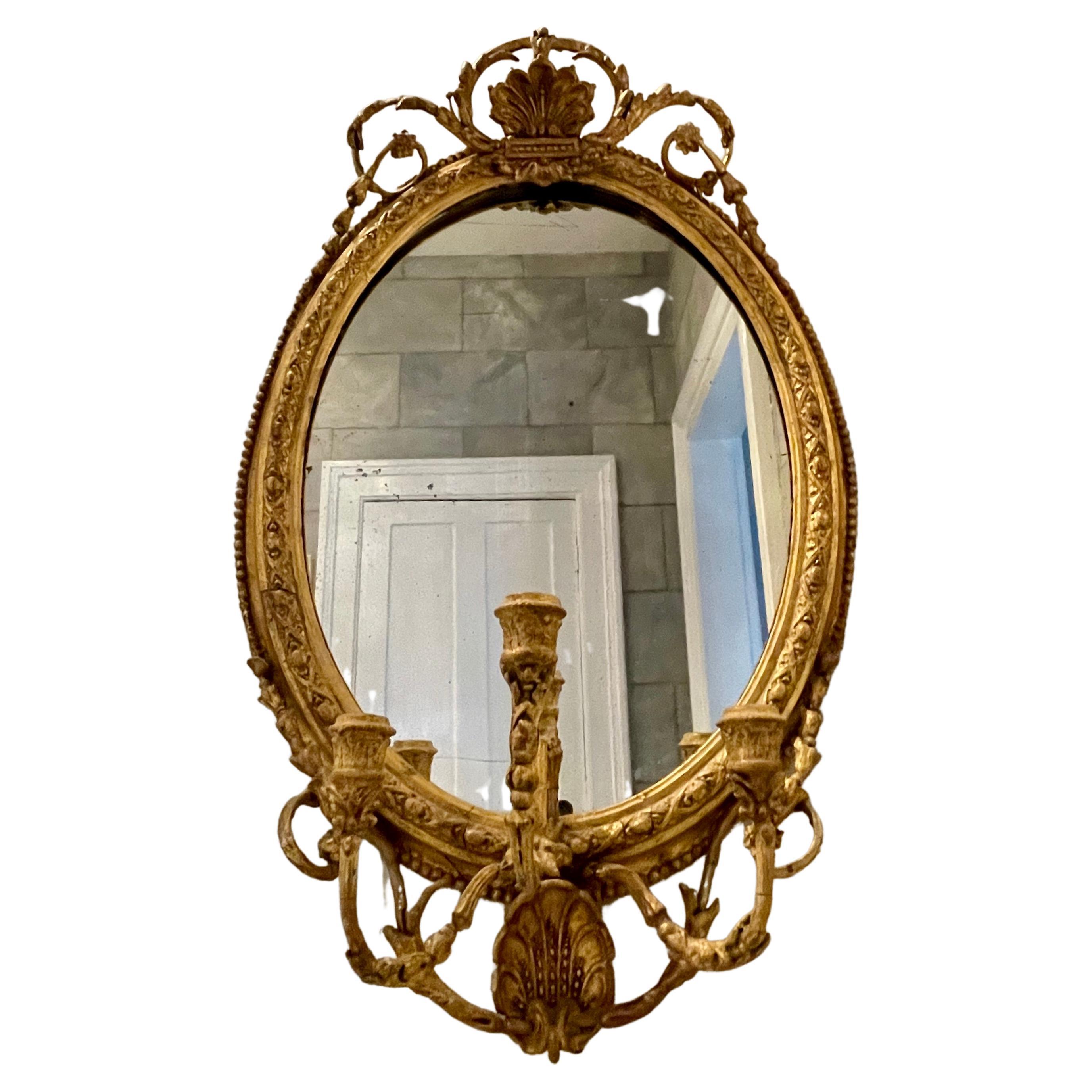 This delicate oval mirror is an outstanding example of fine English or German craftsmanship in the 18th Century. It features hand-carved wood and gesso gilding with triple candelabra, double shell decoration, beaded bellflower molding, and original