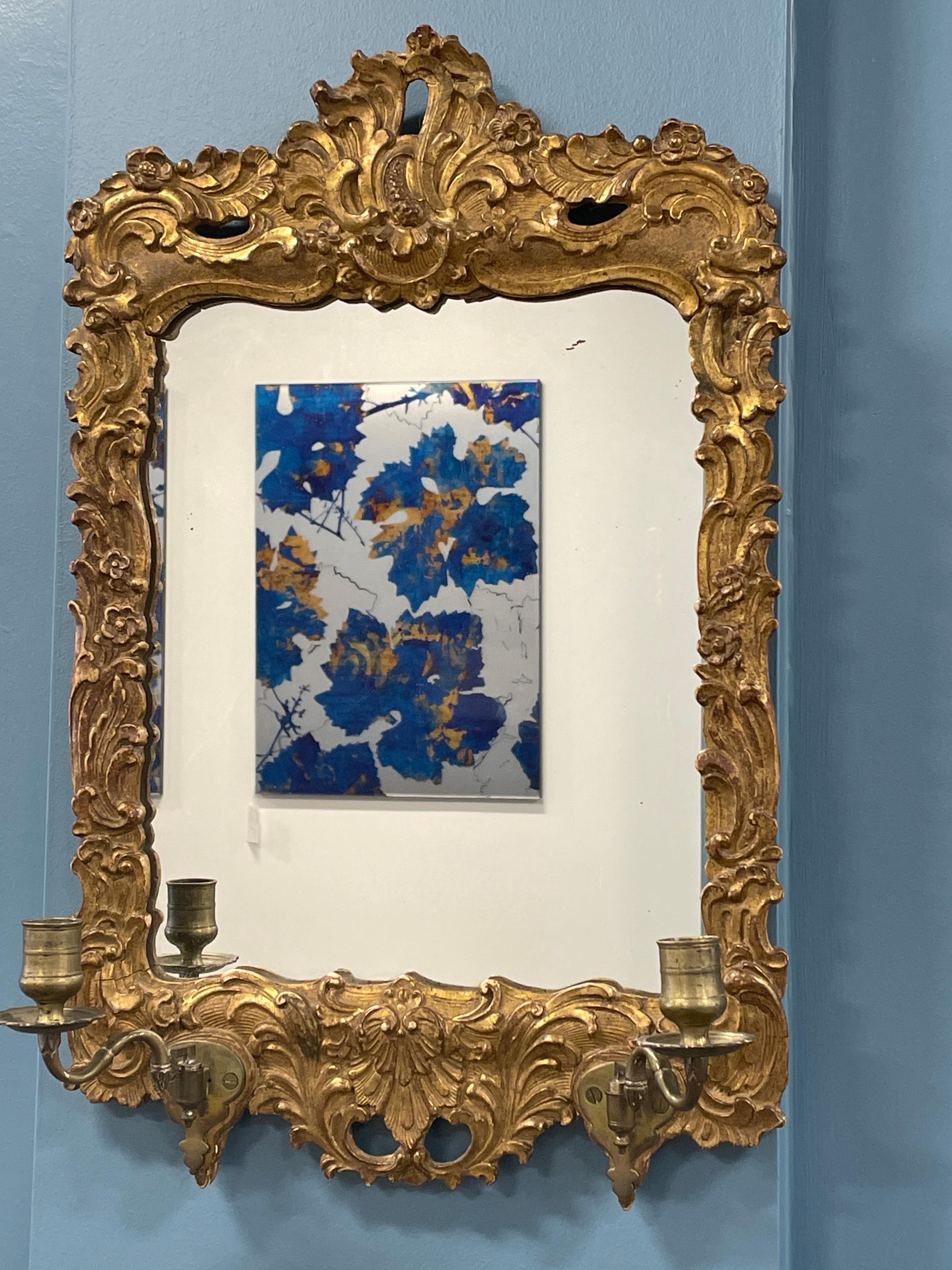 Élevez votre espace avec l'élégance intemporelle de cet authentique miroir baroque allemand orné de chandeliers. Fabriqué en bois doré, chaque détail est minutieusement sculpté à la main avec de délicats motifs de rocaille et de fleurs. Le verre du