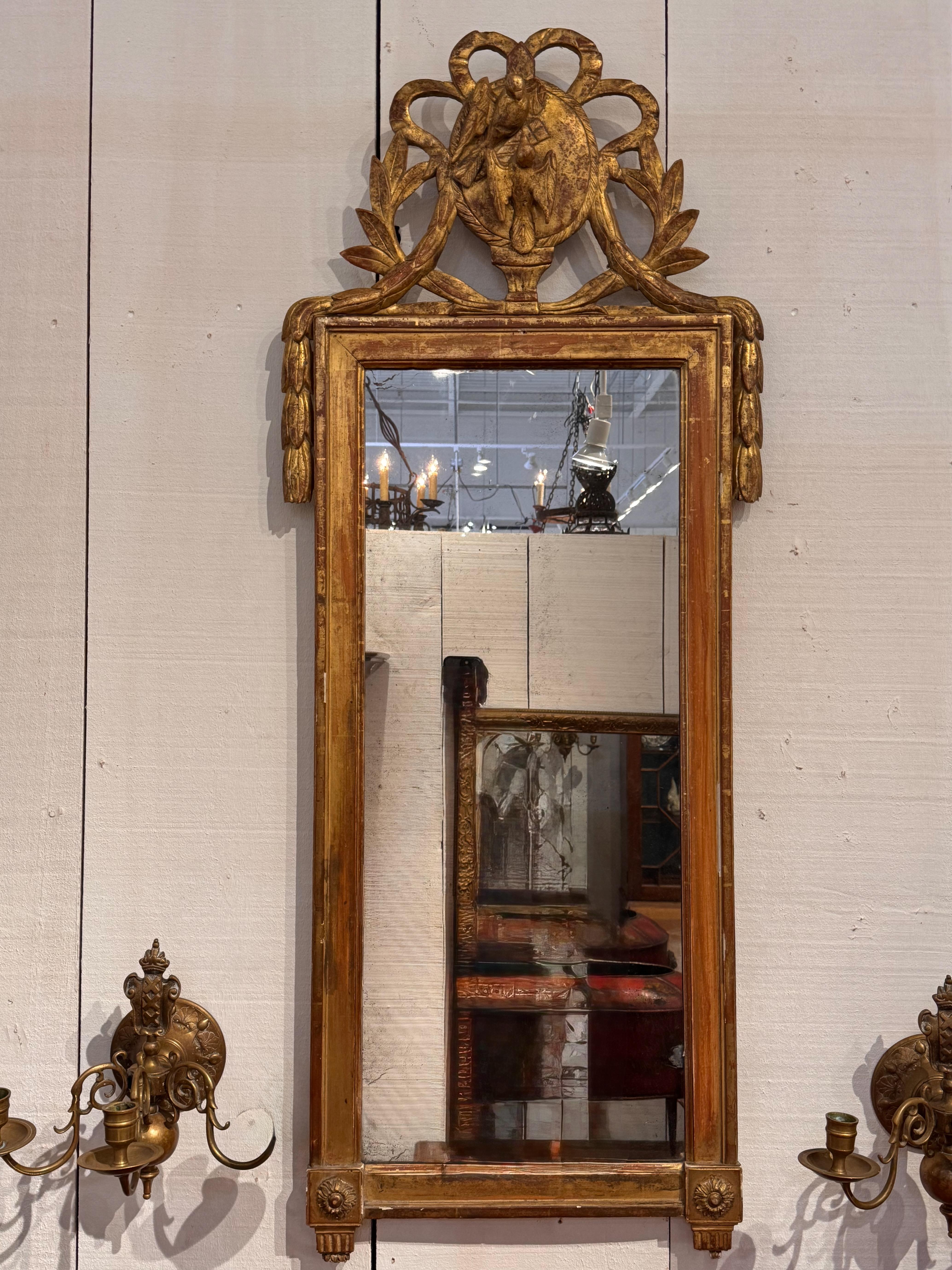 Este espejo de madera dorada del siglo XVIII presenta una cresta finamente tallada con un medallón central que representa una paloma y un polluelo en relieve -emblema de paz y crianza-, enmarcado por ramas de laurel atadas y detalles de cintas. La