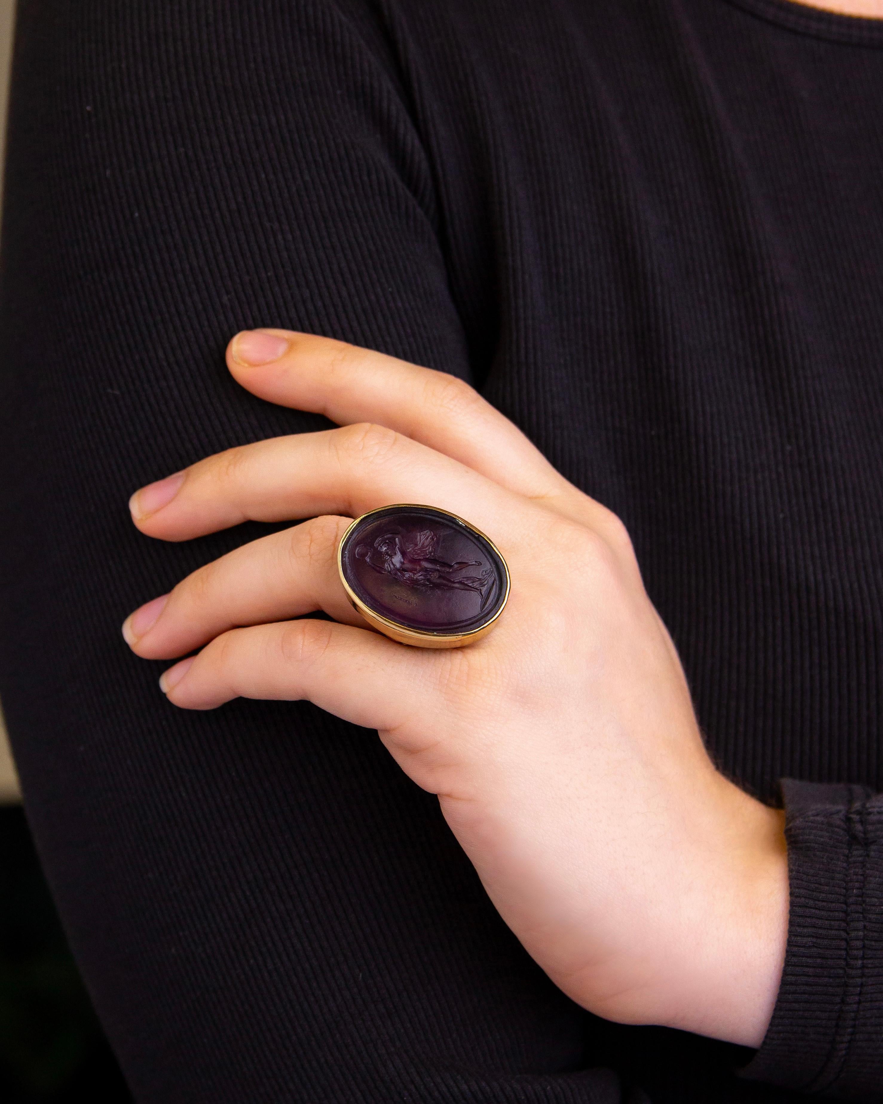 This unusual ring houses what is often referred to as a 'Tassie Glass' intaglio. The aubergine glass intaglio which depicts a nymph with a tambourine likely dates to the late 18th century and has been set in a much later 9 karat yellow gold mount.