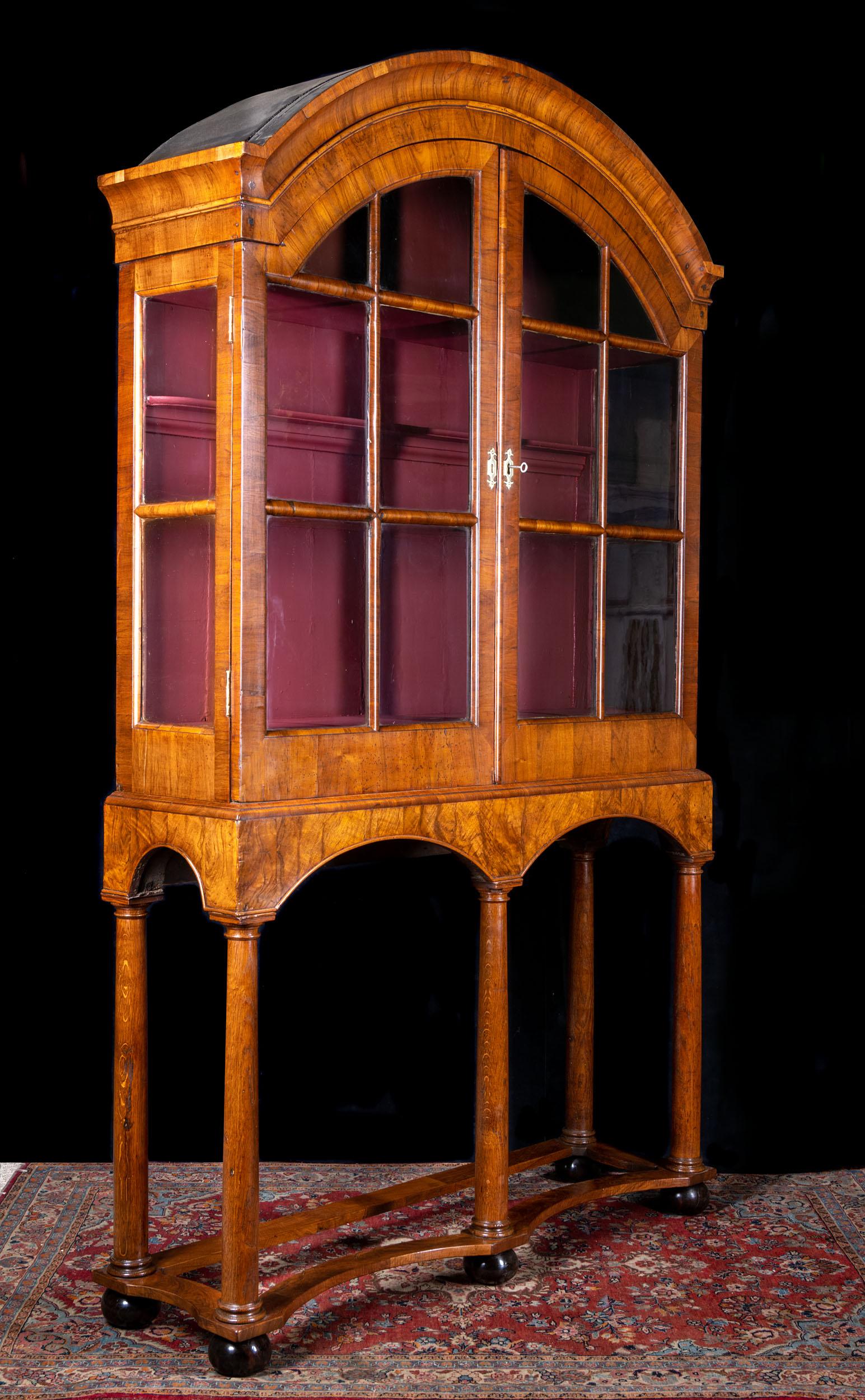 An early 18th century glazed walnut vitrine cabinet on stand. The arch topped display cabinet has its original glass and slender astragals, revealing two shelves and a concealed drawer. The elegant stand of Tuscan column formed legs, are raised on