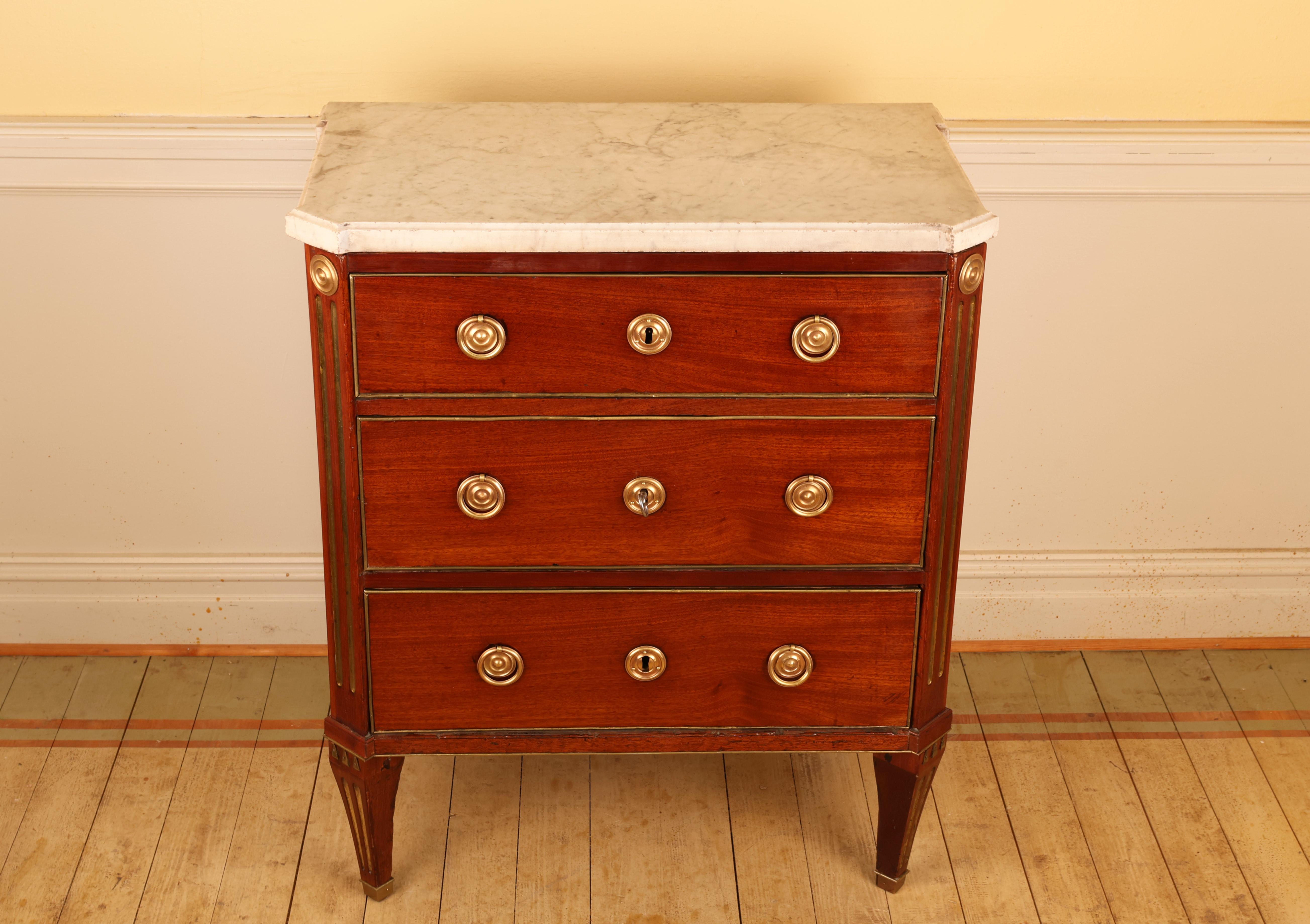 A Gustavian Stockholm piece, dating to the latter part of the 18th century. The commode is veneered in mahogany, with fittings and moldings in brass. It features canted corners adorned with gilded fluting and a marble top. The chest has three