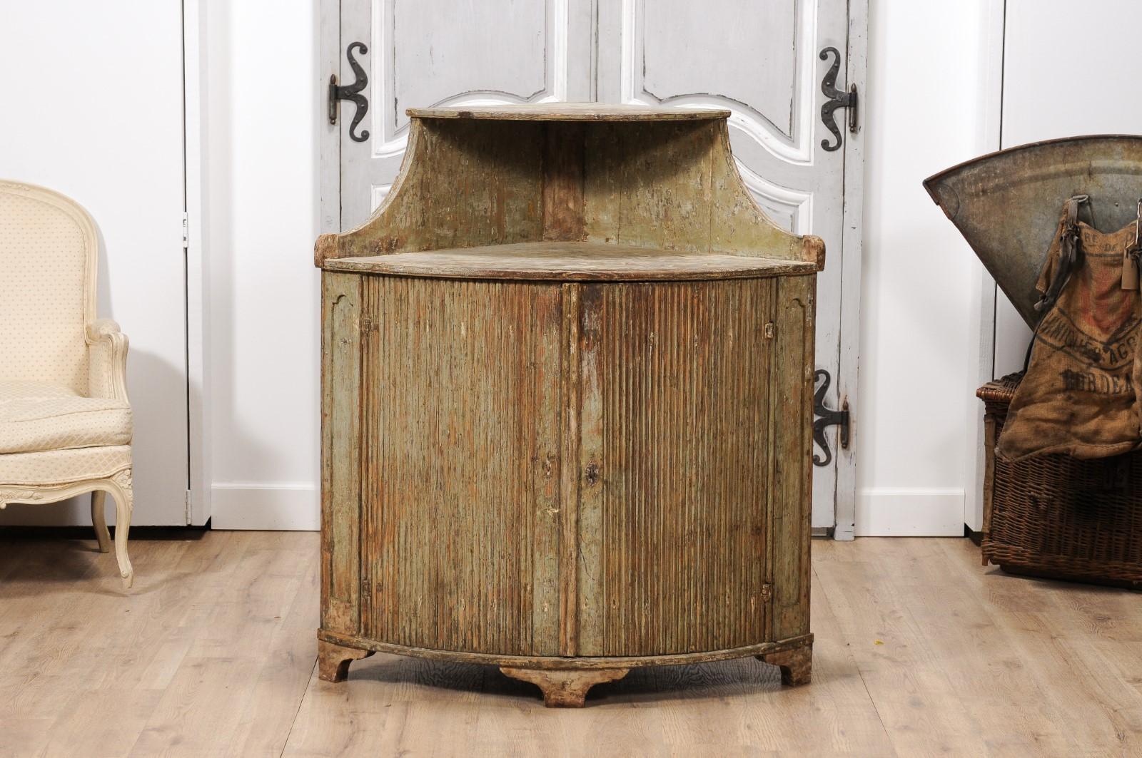 A Swedish Gustavian period painted corner buffet from the 18th century with open shelf at the top, bow front, carved reeded doors, simply carved bracket feet and distressed finish. Immerse yourself in the world of historic Scandinavian elegance with