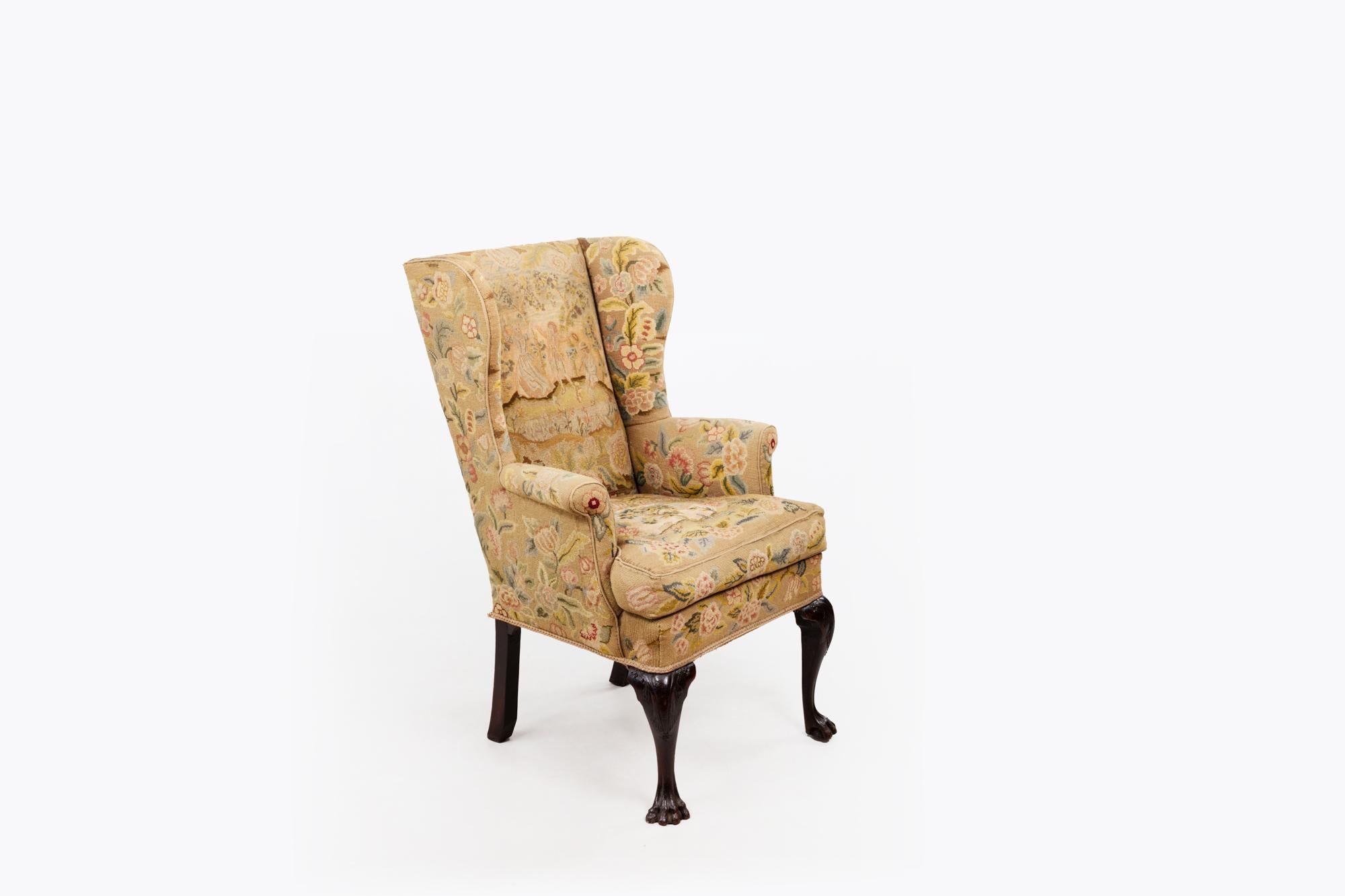 18th Century Irish George II wingback armchair featuring carved cabriole legs terminating in lion paw feet to the front, and simple legs to the back. The chair is covered in needlework upholstery depicting country scenes and floral garlands.