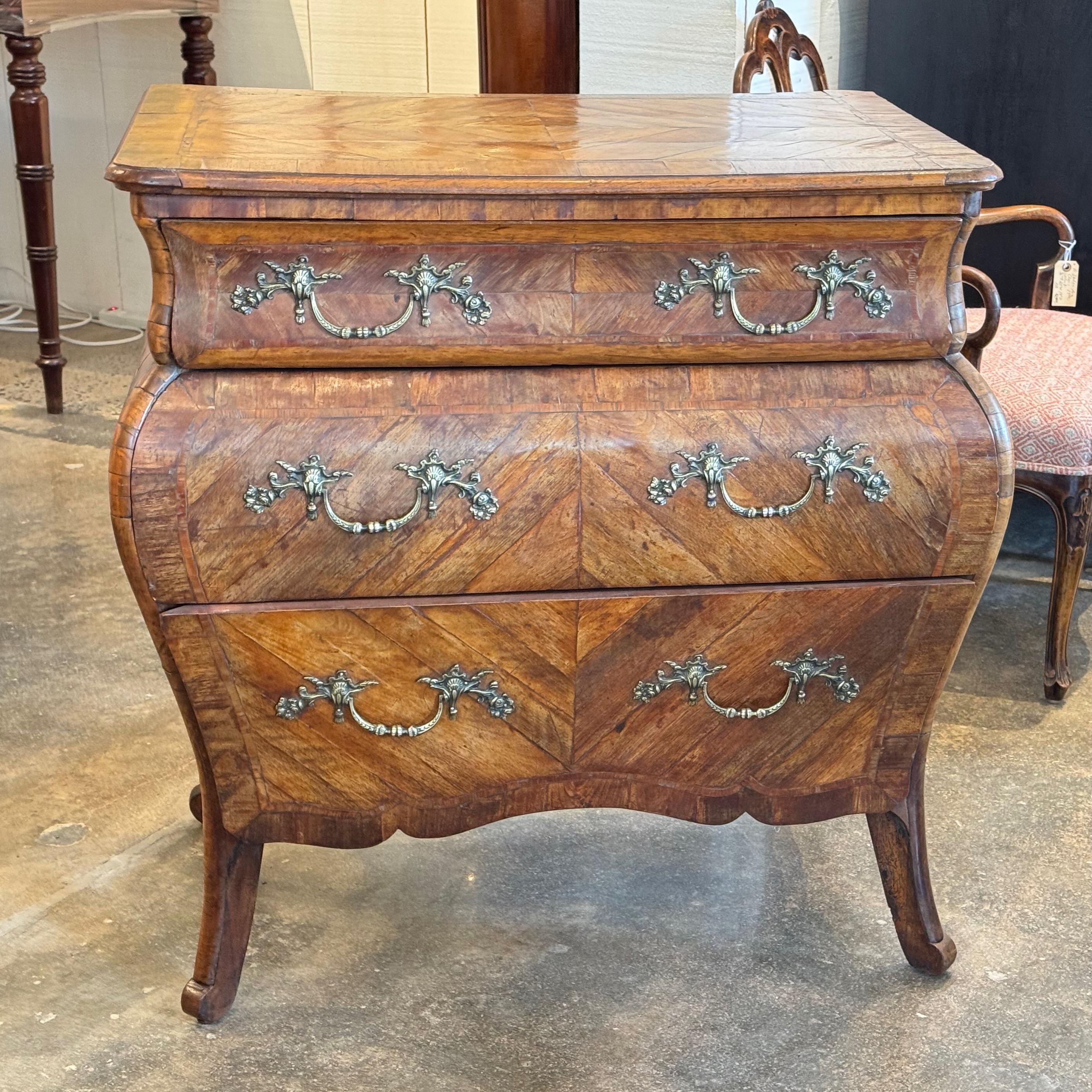 Commode italienne de forme bombée en placage de marqueterie à motif de chevrons. La pièce comprend trois tiroirs avec des poignées en laiton ornées et repose sur des pieds incurvés avec un pied en volute souple, typique de l'influence du baroque