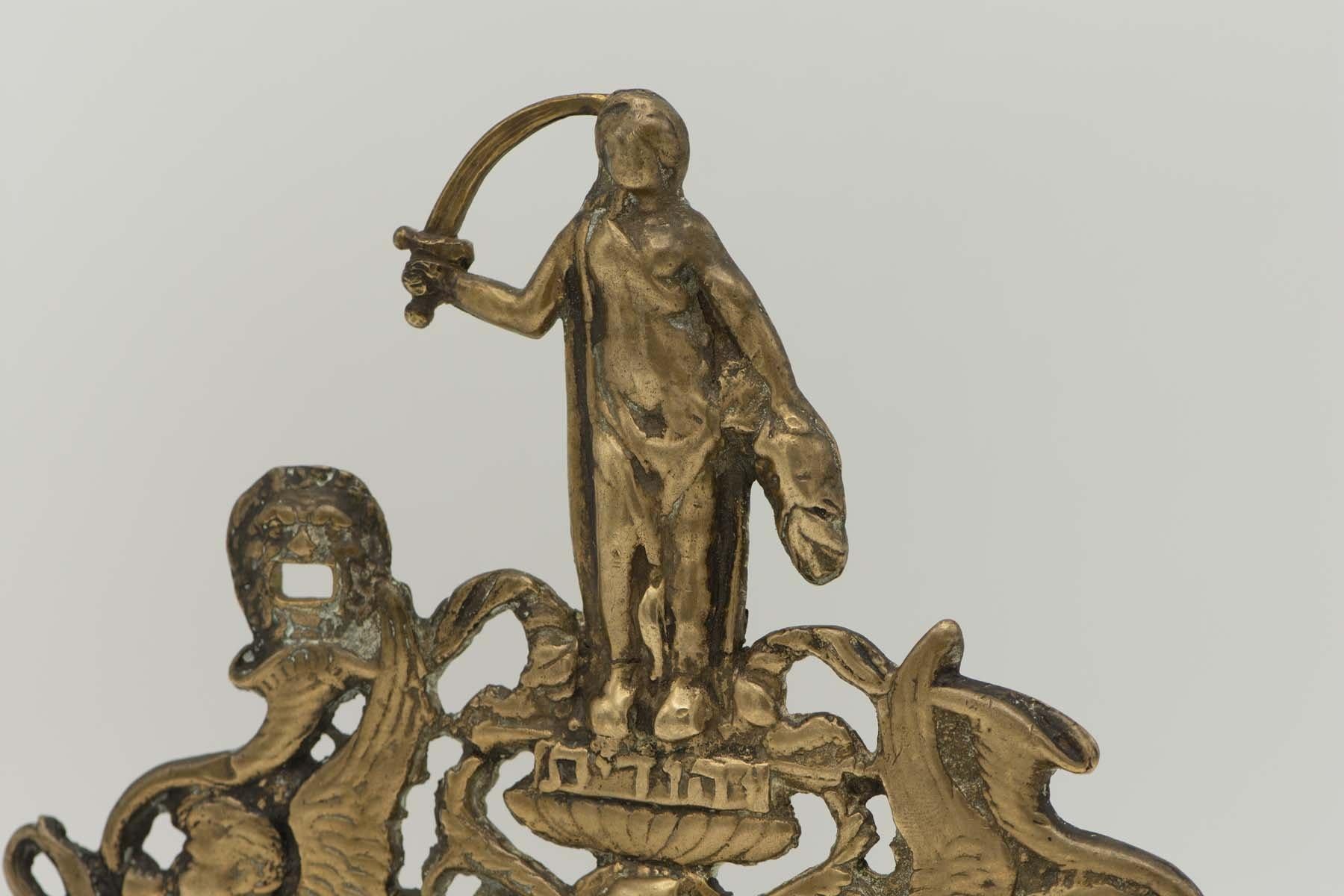 Brass Hanukkah lamp menorah, Italy, circa 1750. 
The backplate is decorated with two winged figures holding vases and blowing trumpets, and backed by a symbol of a bent leg with vase shaped pediment upon which stands the figure of Judith holding a