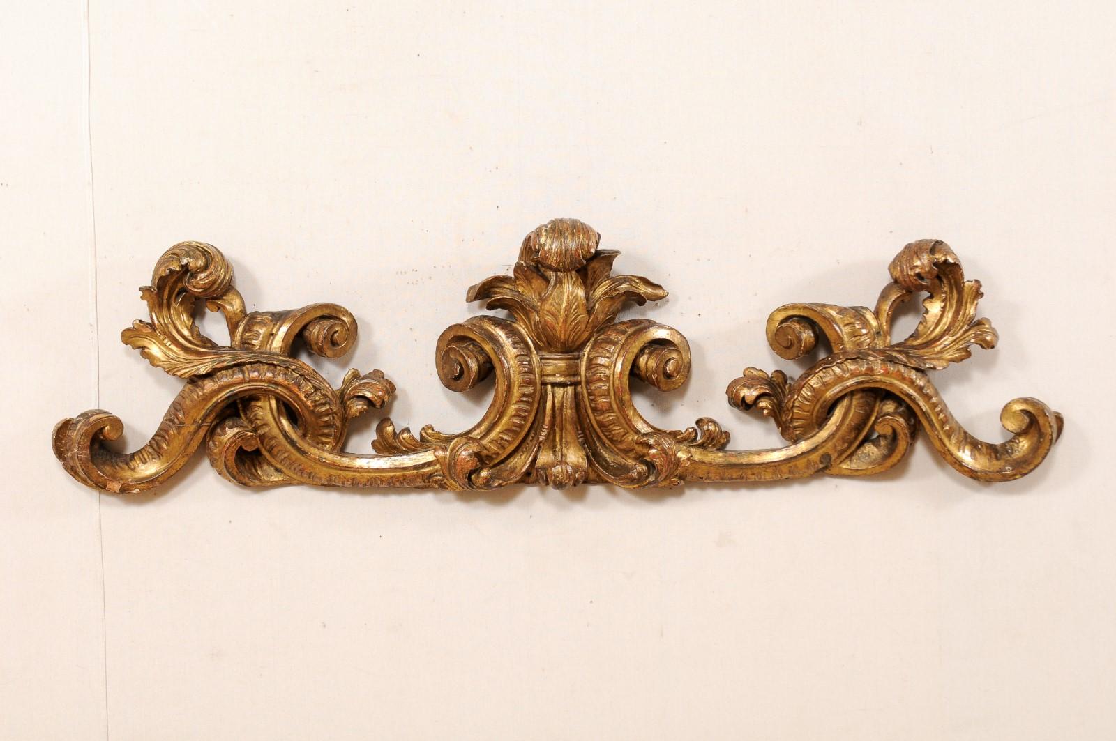 An Italian 18th century gilt and carved wood wall fragment. This antique Italian wall decoration has a drawn out, more horizontally positioned shape, which is heavily carved in a scroll and acanthus leave motif. The top of the plaque features three