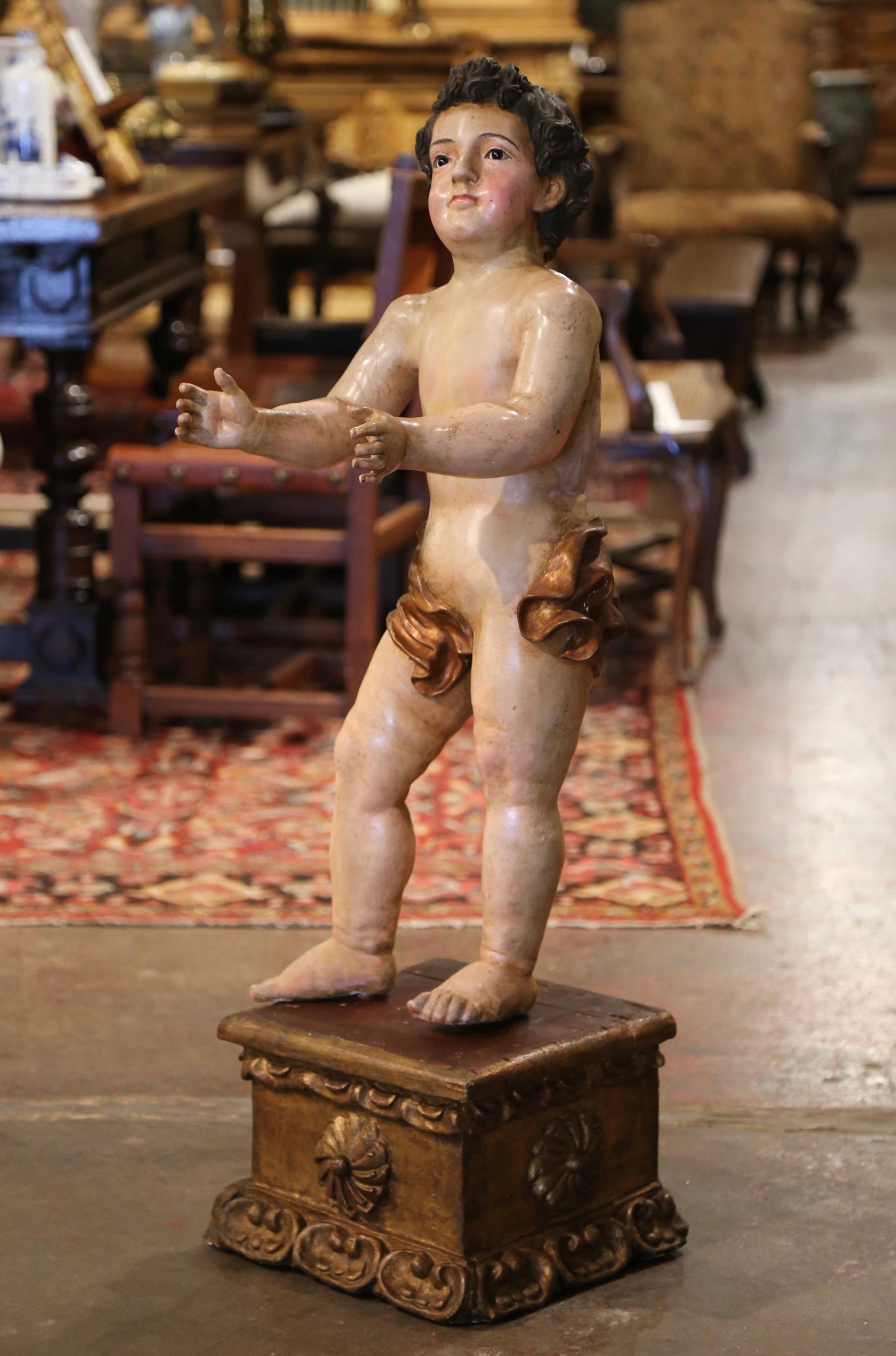 Hand crafted in Italy circa 1780, the antique cherub stands on a square base decorated with shell and foliage motifs. The hand carved sculpture depicts a life size young boy wearing a cloth around his waist, his arms extending and looking up; he is