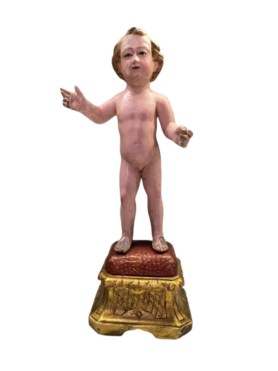 Wir präsentieren eine außergewöhnliche, aus Holz geschnitzte und polychromierte Figur aus dem 18. Jahrhundert, die das zarte Bild des Jesuskindes in stehender Haltung zeigt, das anmutig auf einem kunstvoll beblätterten Kissen und Sockel thront.