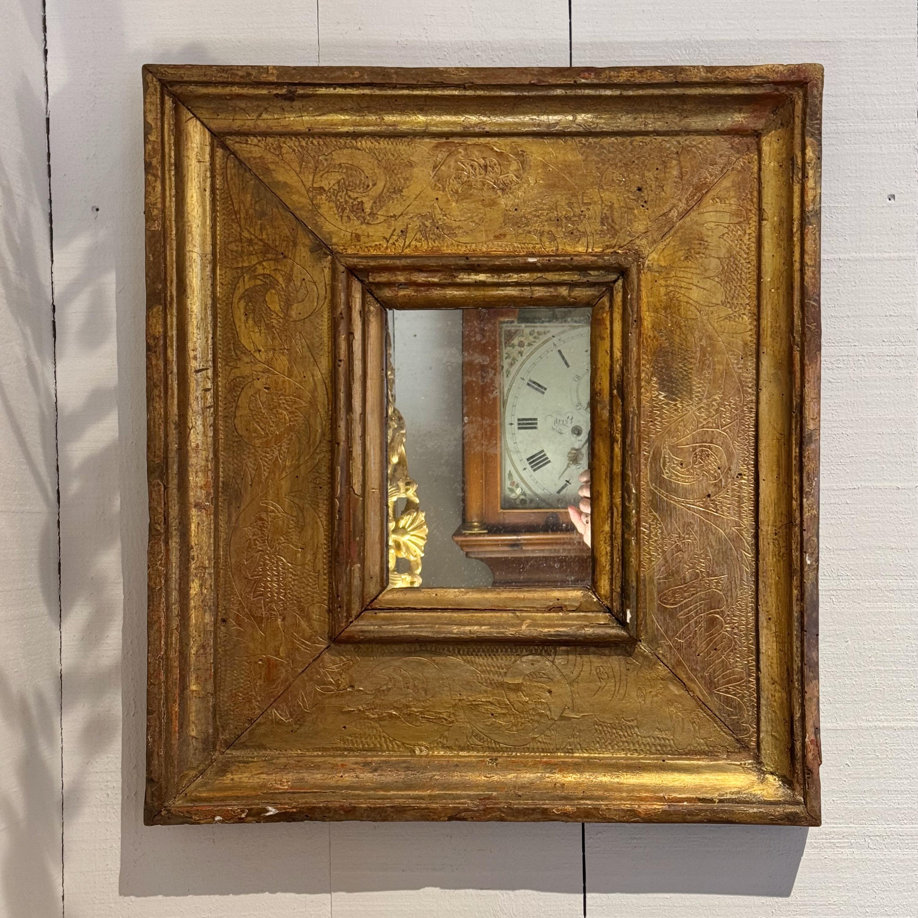 Compacto espejo italiano del siglo XVIII con un marco ancho de madera dorada profundamente biselado. La superficie presenta motivos foliados y volutas incisas, suavizados por la edad y una rica pátina. La moldura interior añade dimensión y enmarca