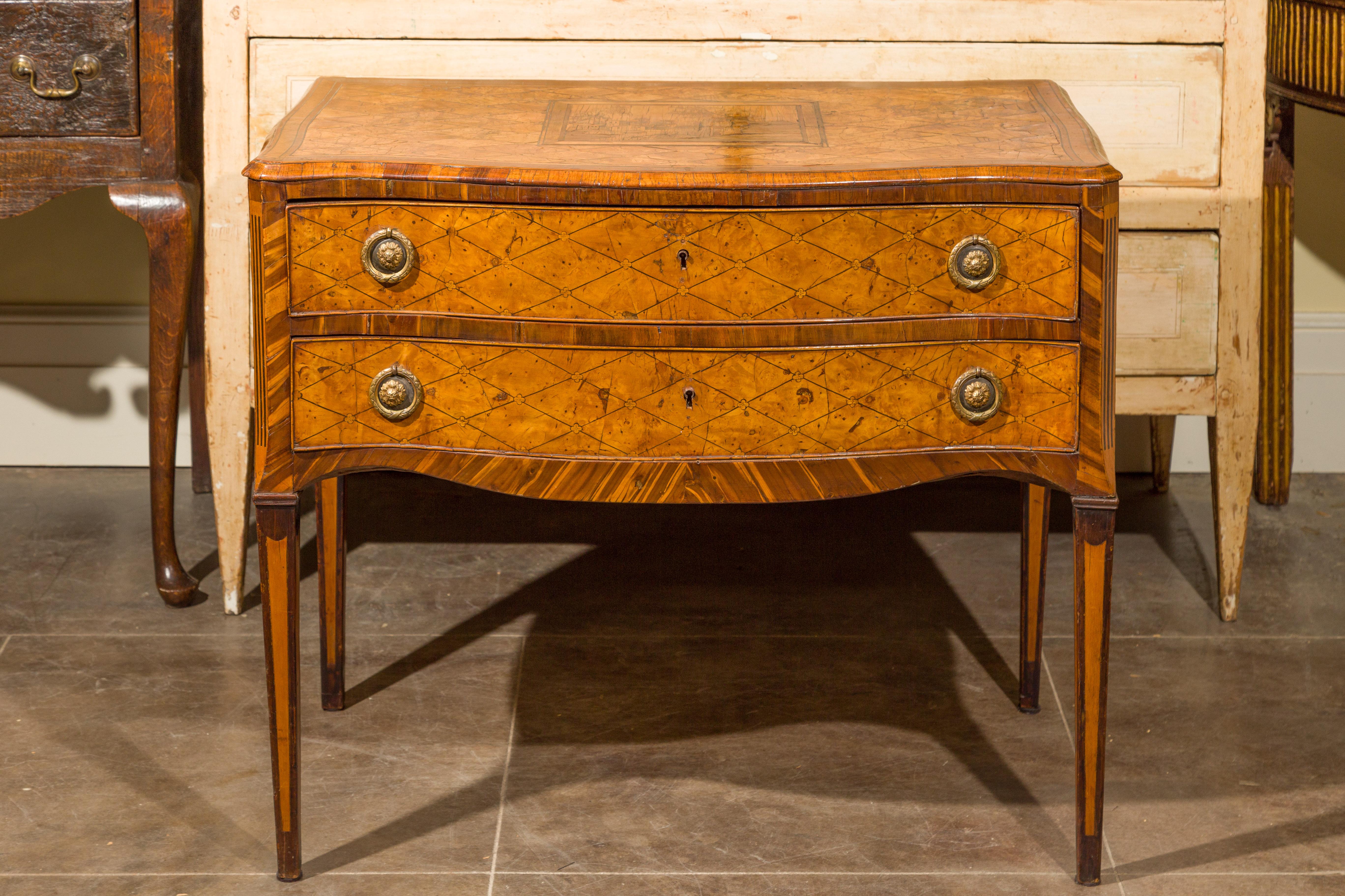 Esta cómoda italiana del siglo XVIII presenta una exquisita tapa adornada con una chapa de madera con motivos de rombos, alrededor de una escena paisajística central en marquetería. El motivo de rombos horizontales se repite tanto en los laterales