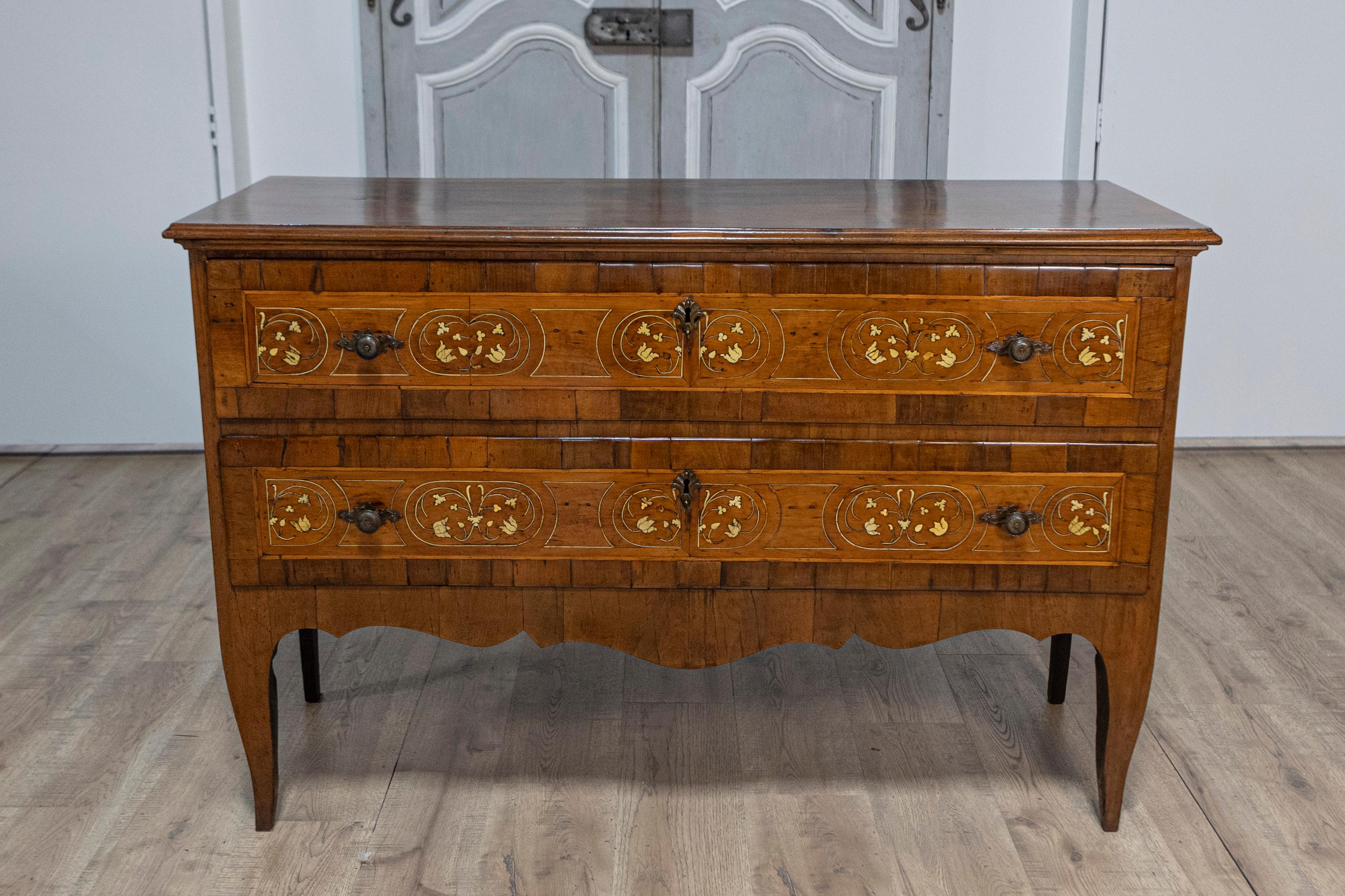 An Italian walnut, mahogany and ash commode from the 18th century with scrollwork marquetry, two drawers and scalloped apron. This exquisite Italian commode from the 18th century is a masterpiece of craftsmanship, featuring a harmonious blend of