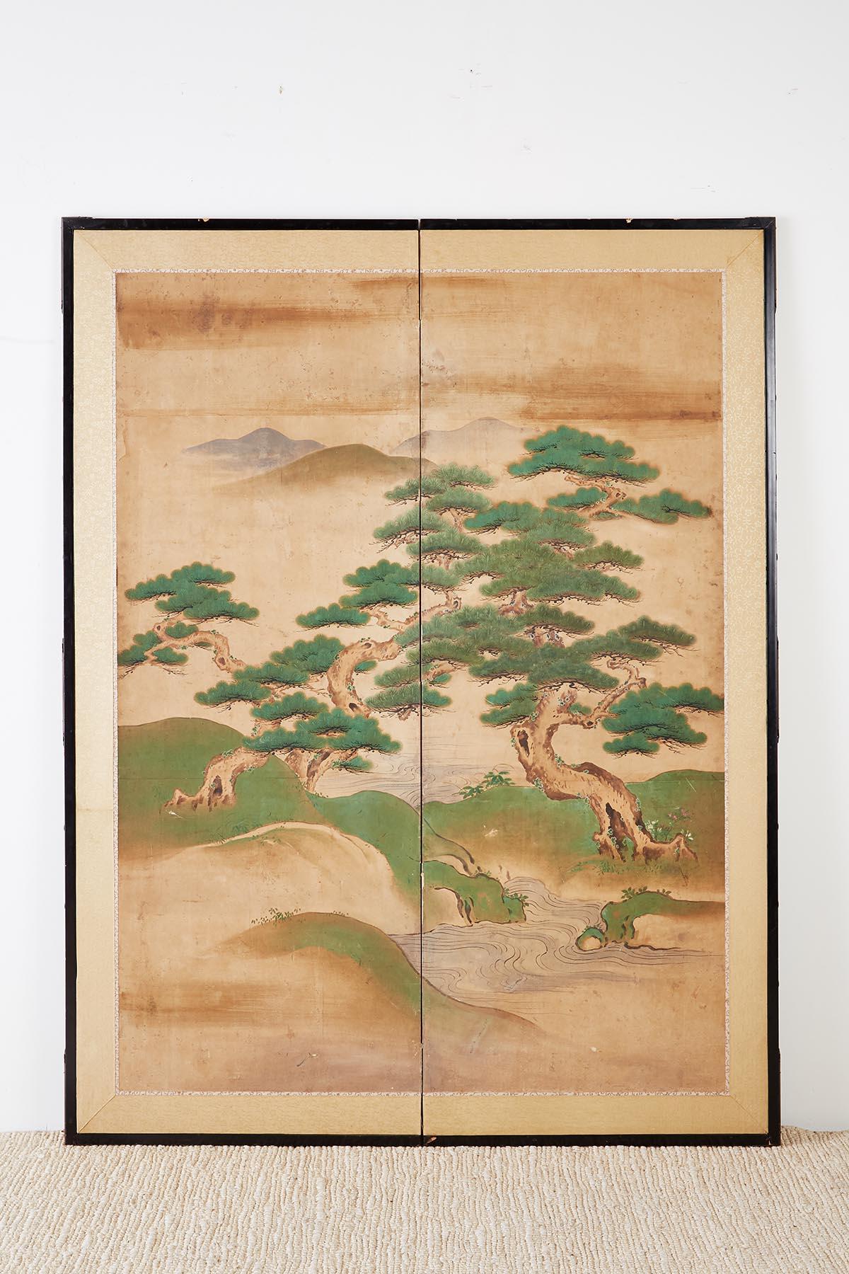 Large late 18th century Japanese two-panel Kano school screen from Edo period. Two large pine trees over a river with mountains in the background. Set with a silk brocade border in an ebonized wood frame. Minor wear and staining on front with losses