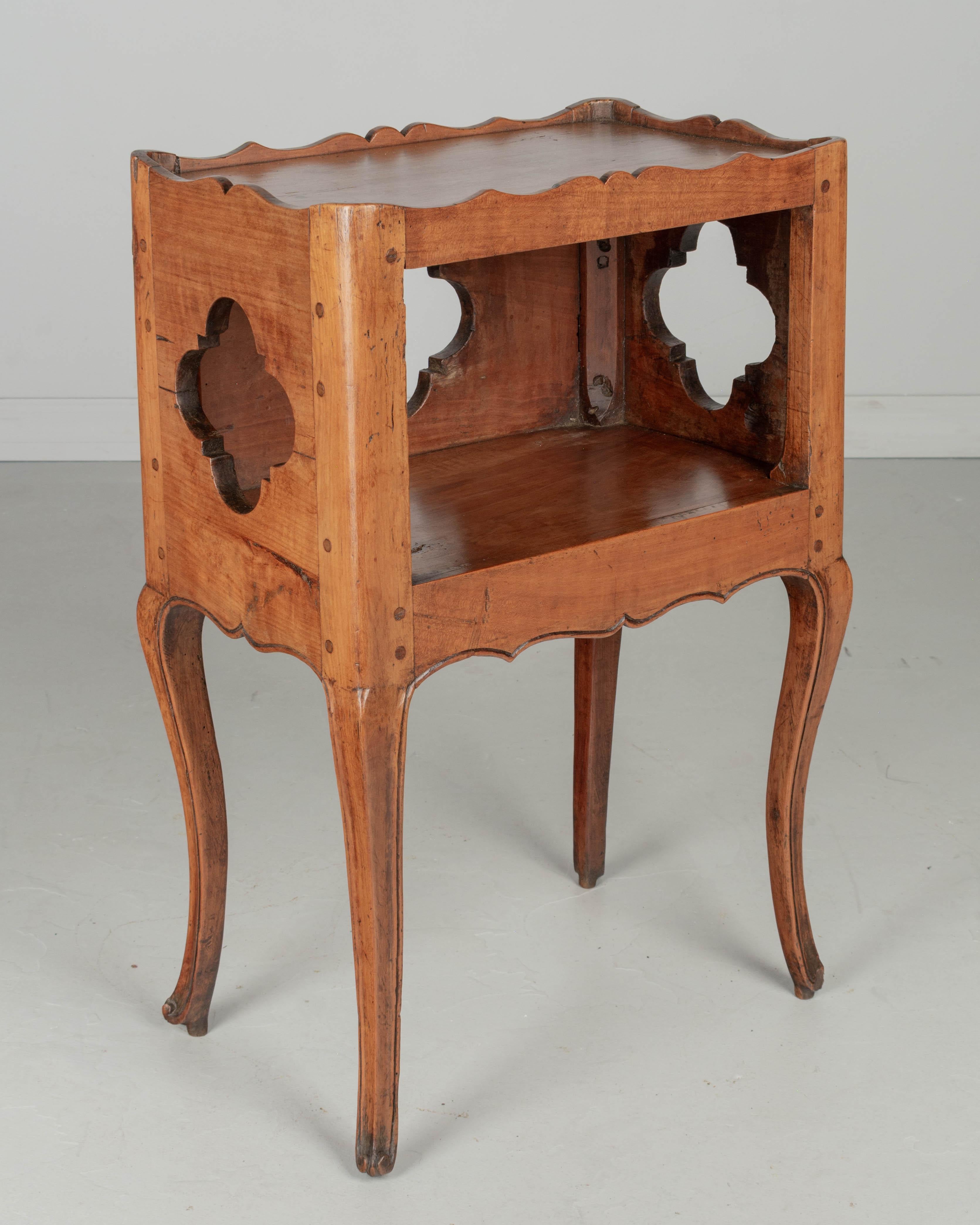 An 18th century Louis XV Country French side table or nightstand made of solid cherry, with open niche and pierced quatrefoil shape cut-outs on the sides and back. Small dovetailed side drawer. Scalloped apron and gallery. Slender curved legs ending
