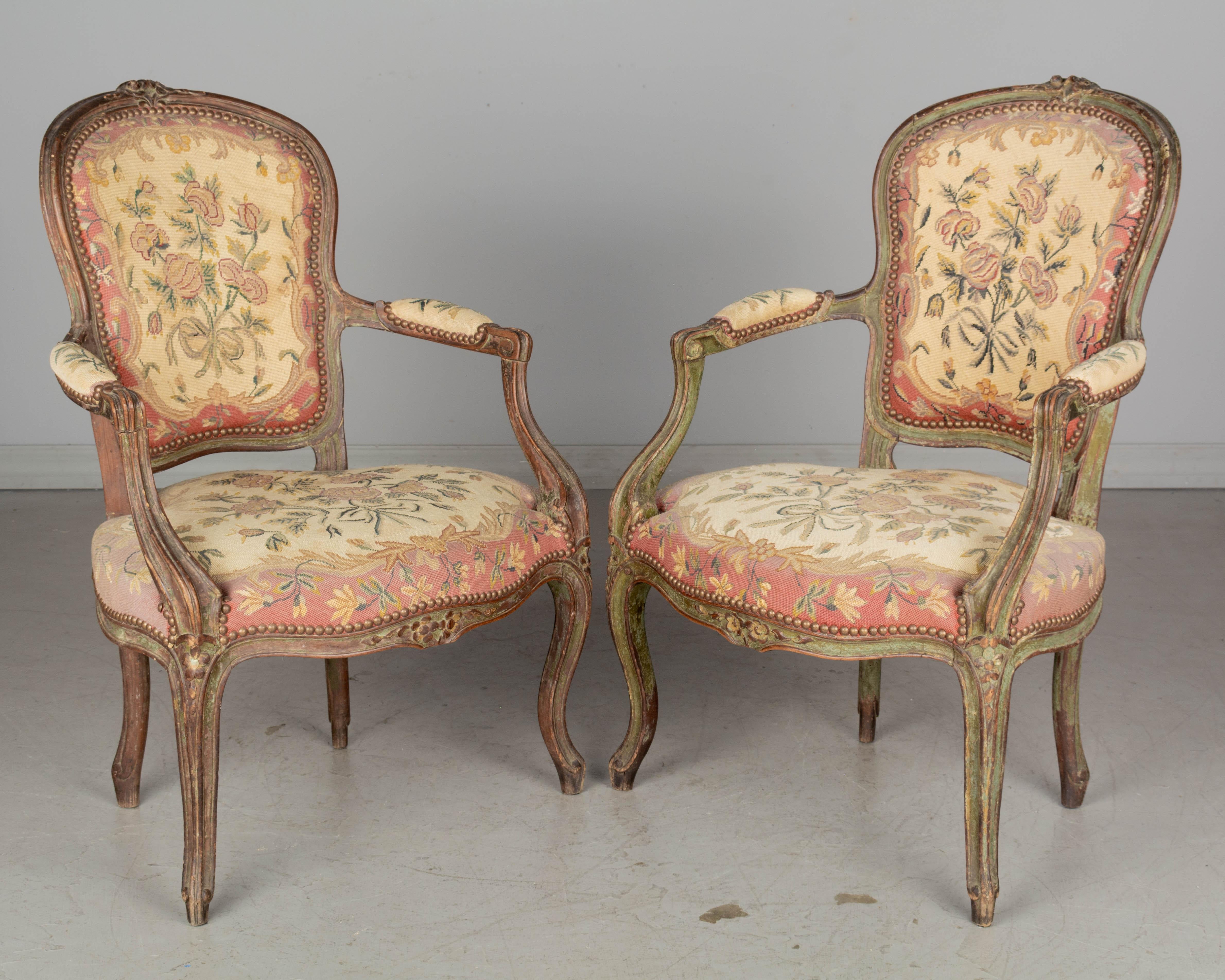A pair of  18th century Louis XV French fauteuils, or armchairs, each with sturdy polychrome painted beechwood frames with hand-carved floral decoration. Upholstered in 19th century petit point tapestry. Pegged construction. Chairs sit low to the