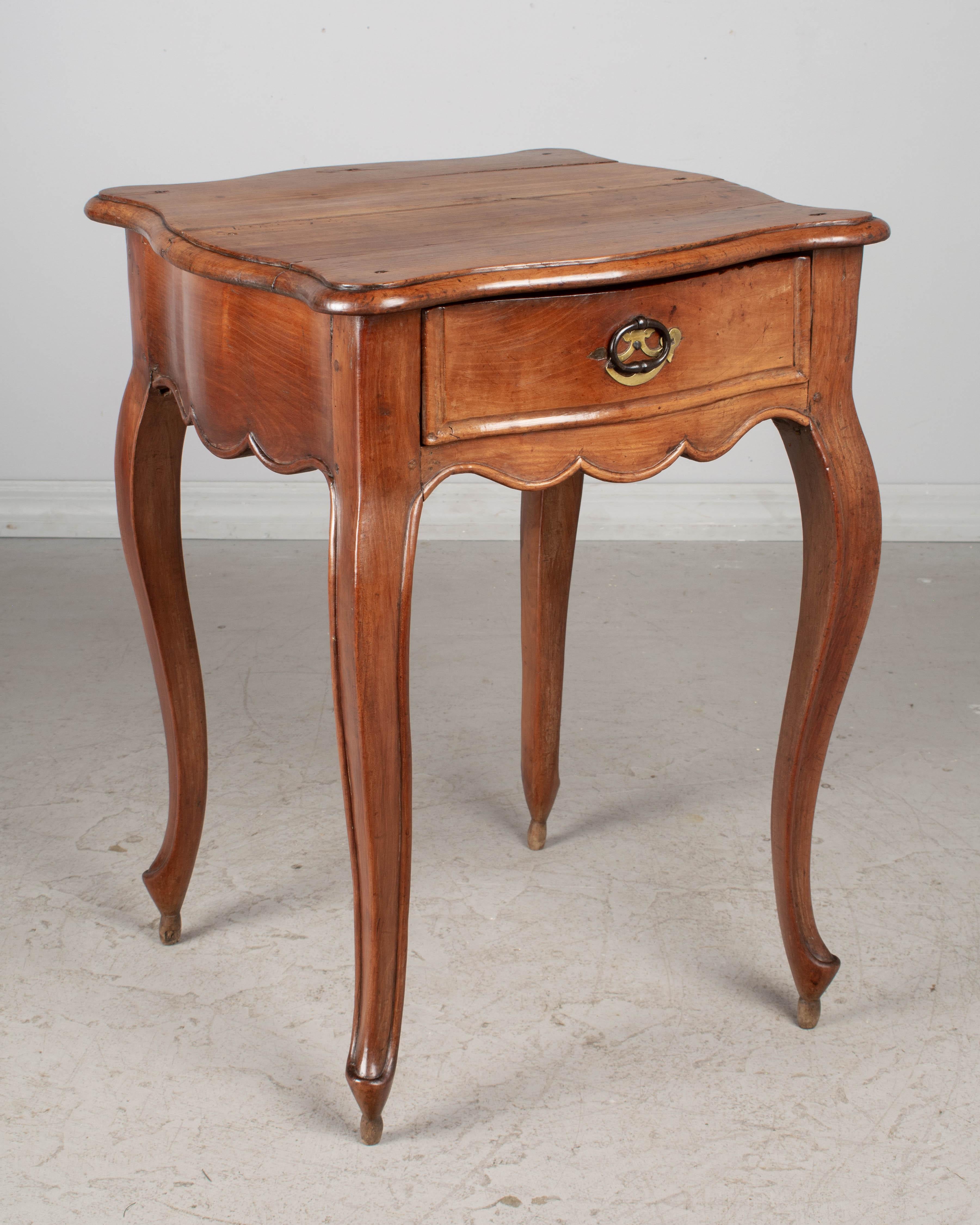 A late 18th century French Louis XV side table from Normandy, made of solid cherry. Rare, elegant serpentine form with shaped top, thick scalloped apron and cabriole legs. Dovetailed drawer one with iron loop pull. Finished on all four sides.