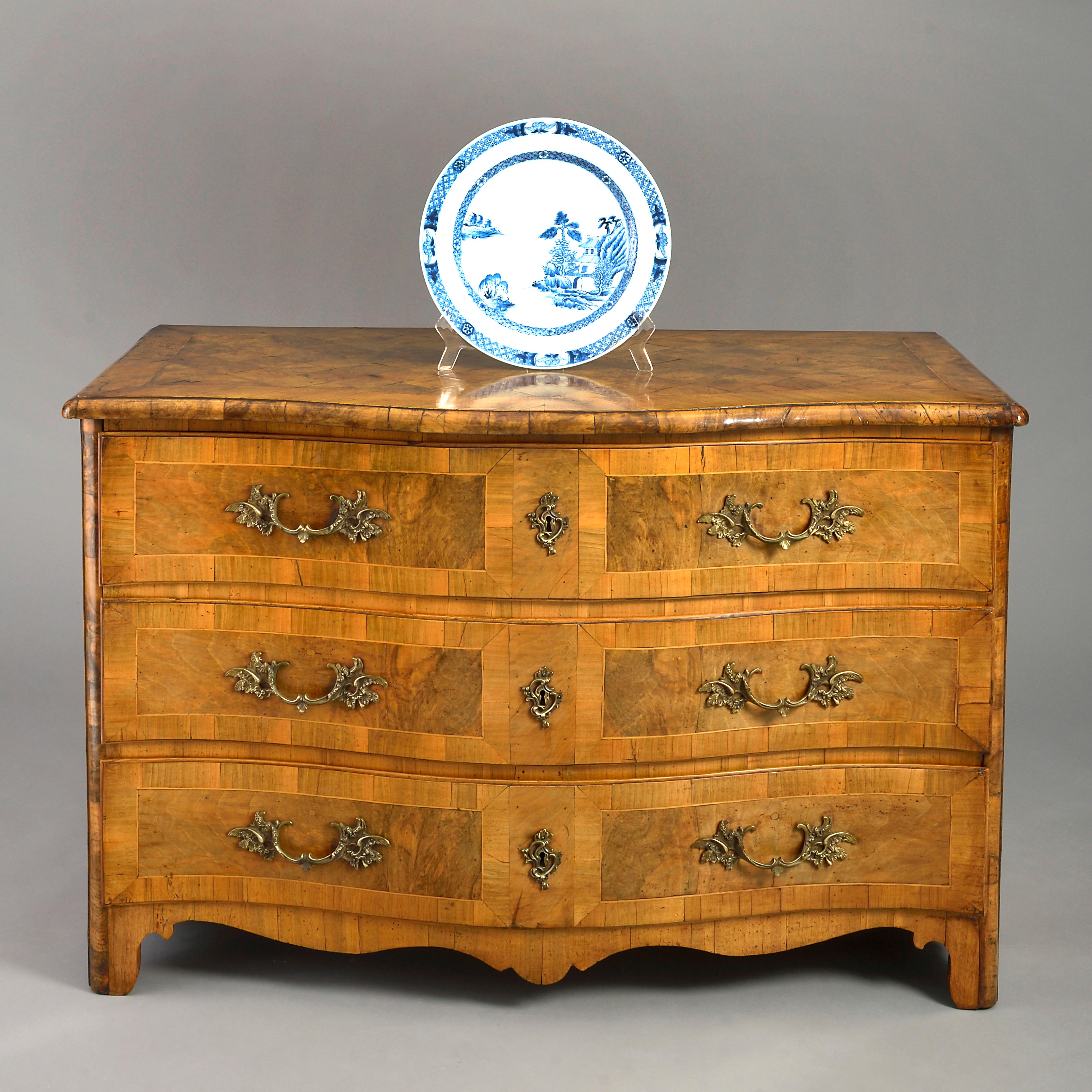 An 18th century Louis XV period commode of serpentine form, the overhanging parquetry top with crossbanding above three long paneled drawers with brass Rococo handles and lock escutcheons and terminating in a shaped apron raised upon bracket feet.