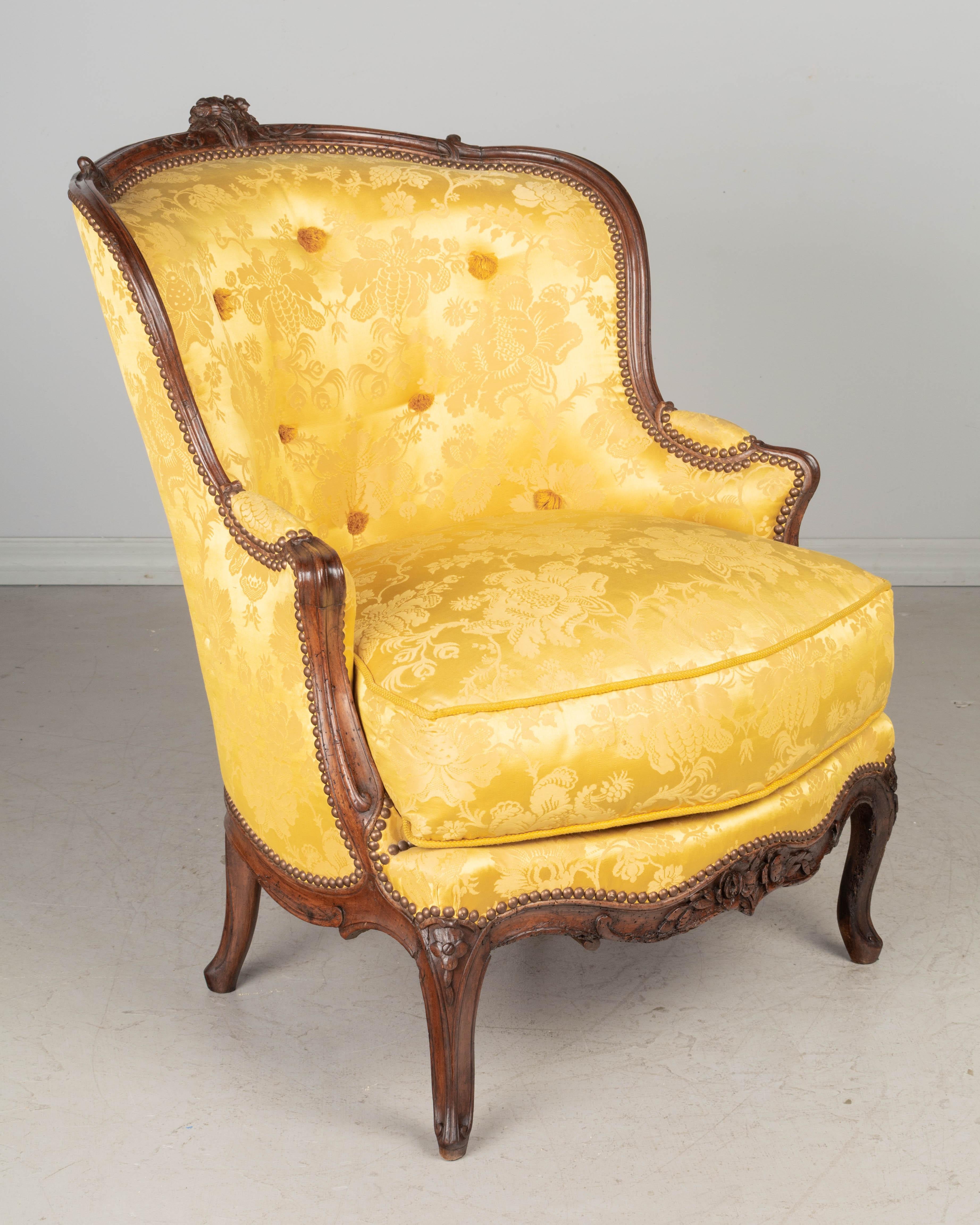 An 18th century French Louis XV bergère or armchair. Sturdy walnut frame with nice hand-carved floral details and crafted using pegged construction. Expertly reupholstered, as found, in bright yellow silk damask fabric with tufting and nailhead