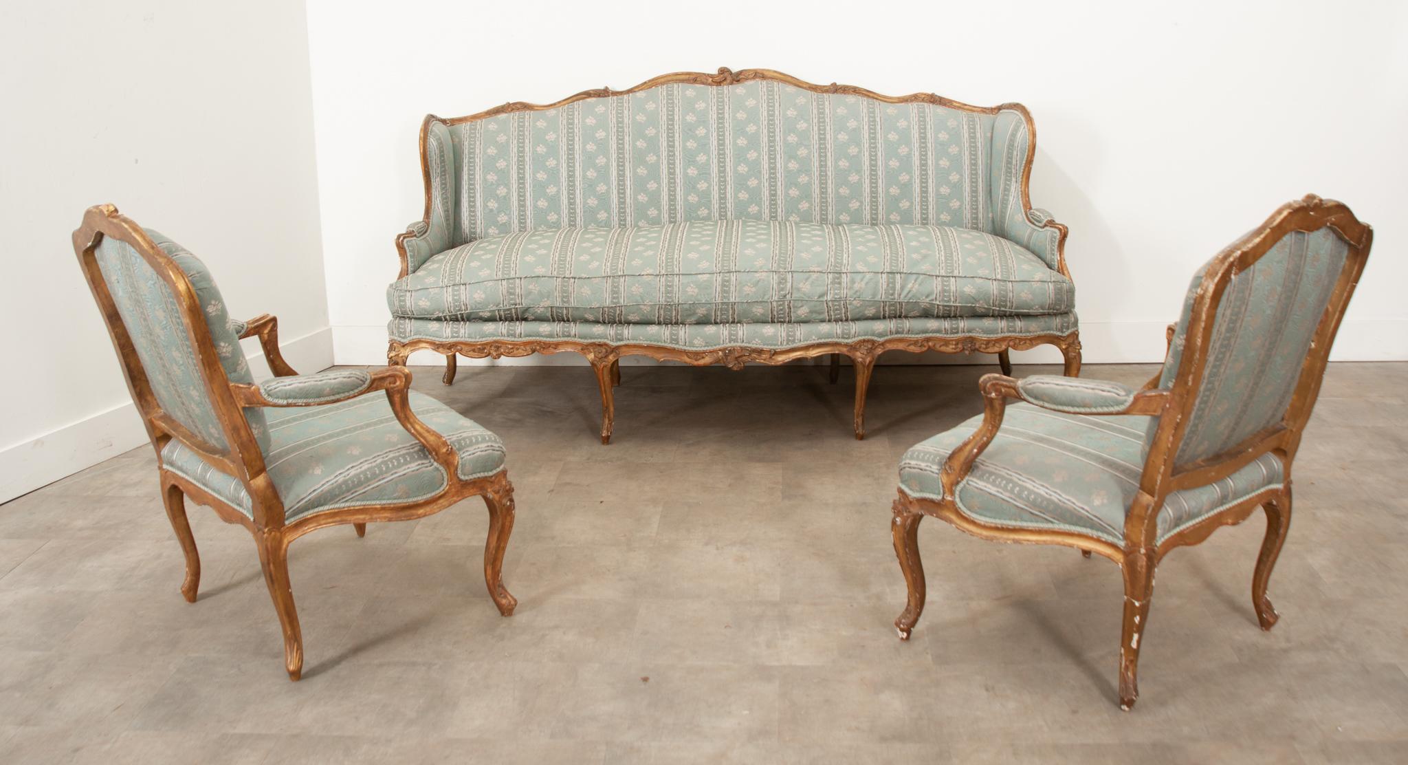 An early 19th century French Louis XV style gold giltwood 3-piece parlor salon suite. This lovely set includes a settee and two armchairs. All pieces feature vintage moire silk upholstery in a stunning pattern with florals and stripes, ribbon trim,