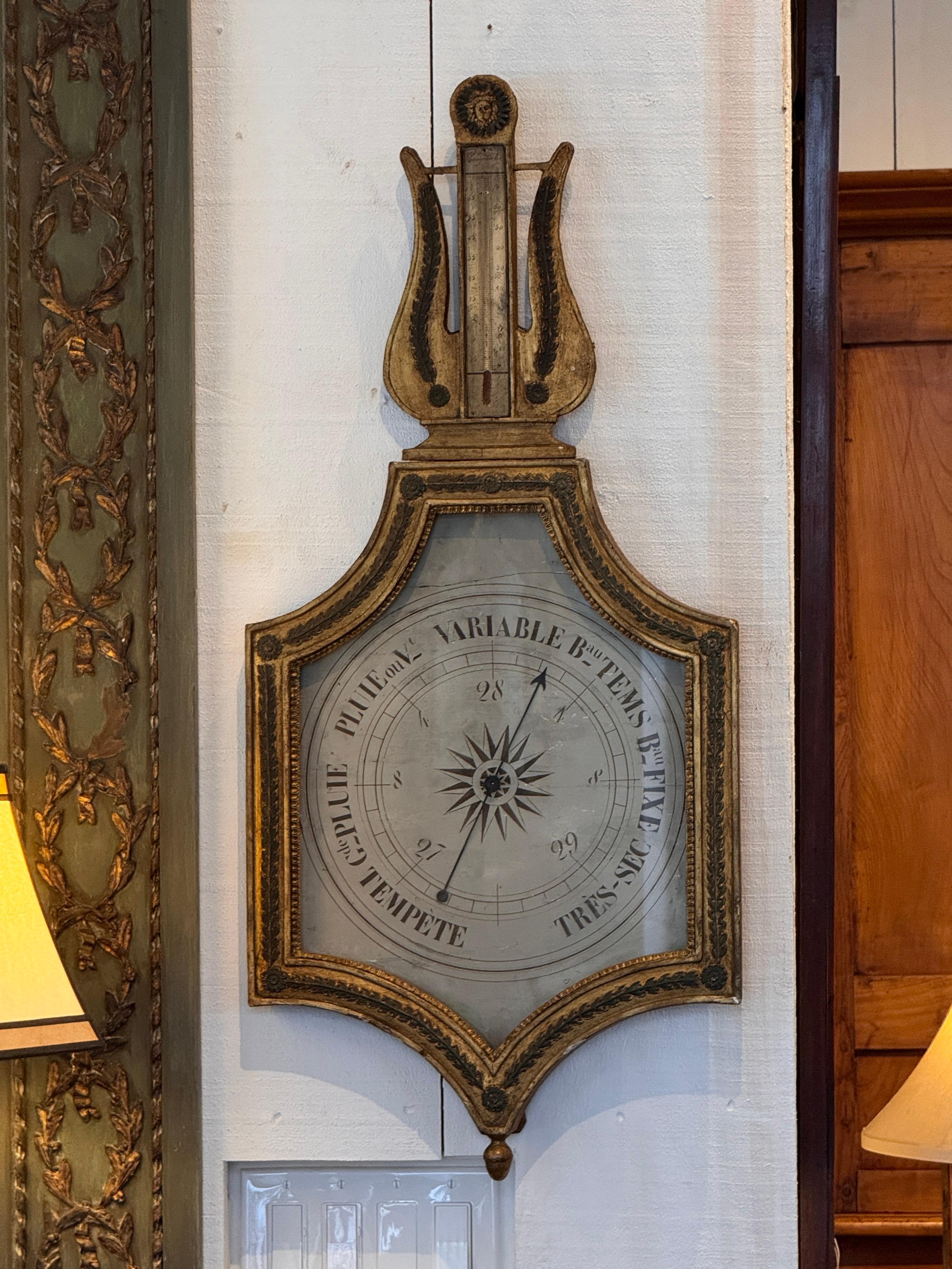 Louis XVI–style giltwood barometer with a lyre-form crest, carved laurel and floral details, and an enameled circular dial marked with French weather indications. The frame shows applied carved ornament and retains an aged gilded surface. A
