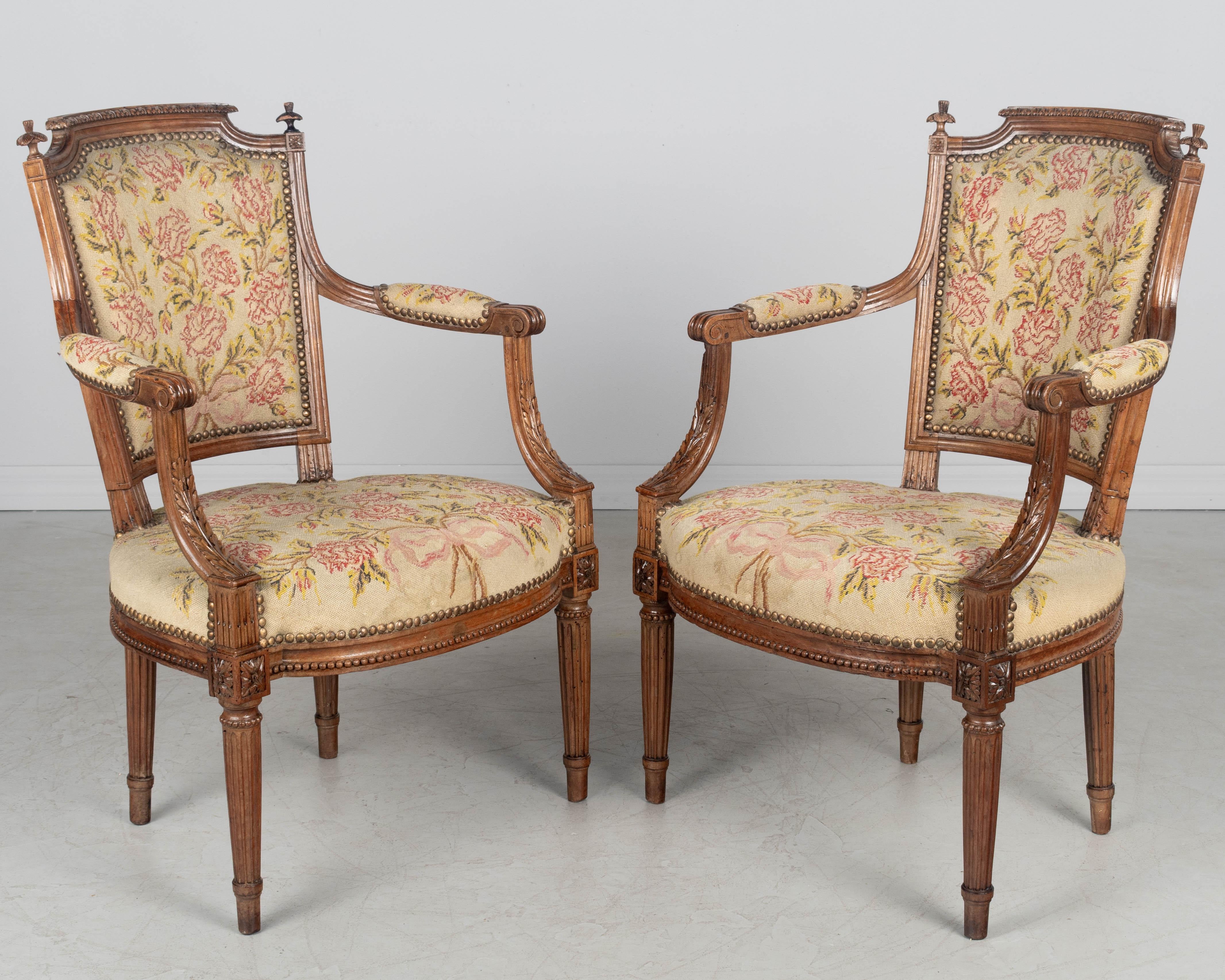 A pair of late 18th Century Louis XVI Period French fauteuils or arm chairs, each with solid carved walnut frame and wool petit point upholstery. Fine carved foliate details with finials on seat back. Sturdy tapered fluted legs. Petite point