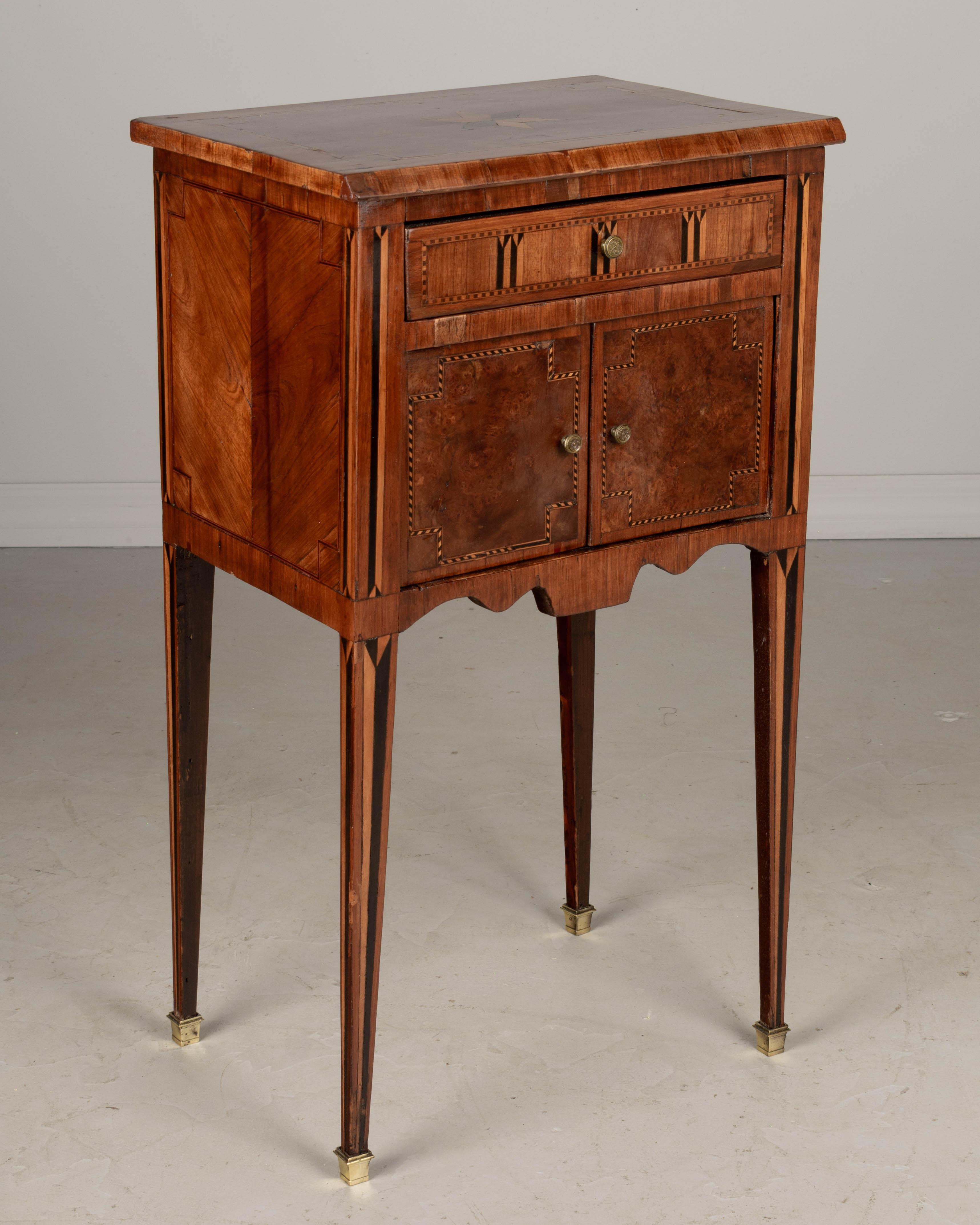 A late 18th century French Louis XVI style marquetry side table, or nightstand, with one drawer above cabinet doors. Made of inlaid veneers of mahogany, walnut and elmwood. Slender tapered legs with marquetry detail and brass sabots. Good