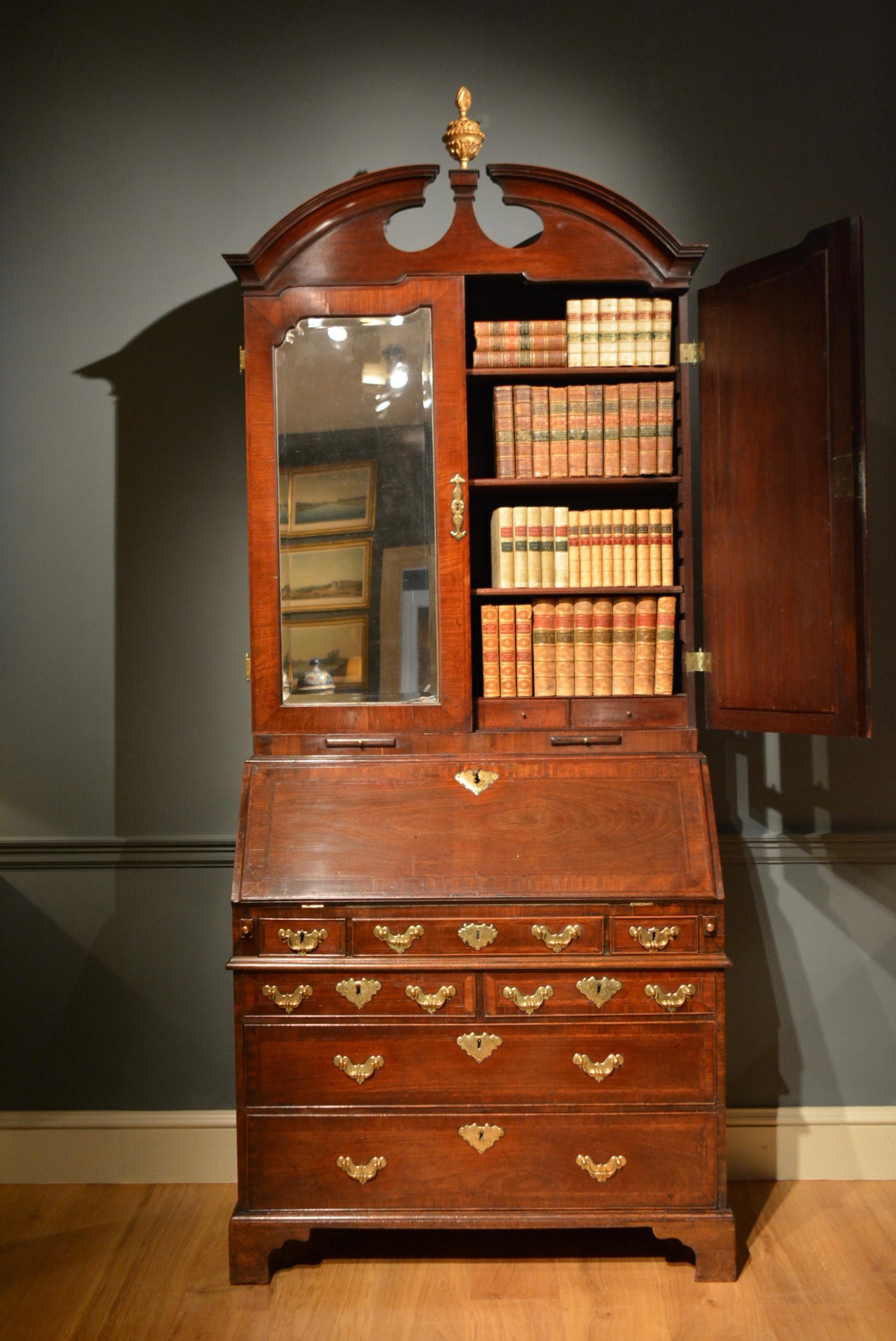 A George II mahogany bureau bookcase, the strongly arched broken pediment centred by a carved gilt finial above a pair of mirrored doors, enclosing adjustable shelves above a bank of small drawers. The fall below opening to reveal a well fitted