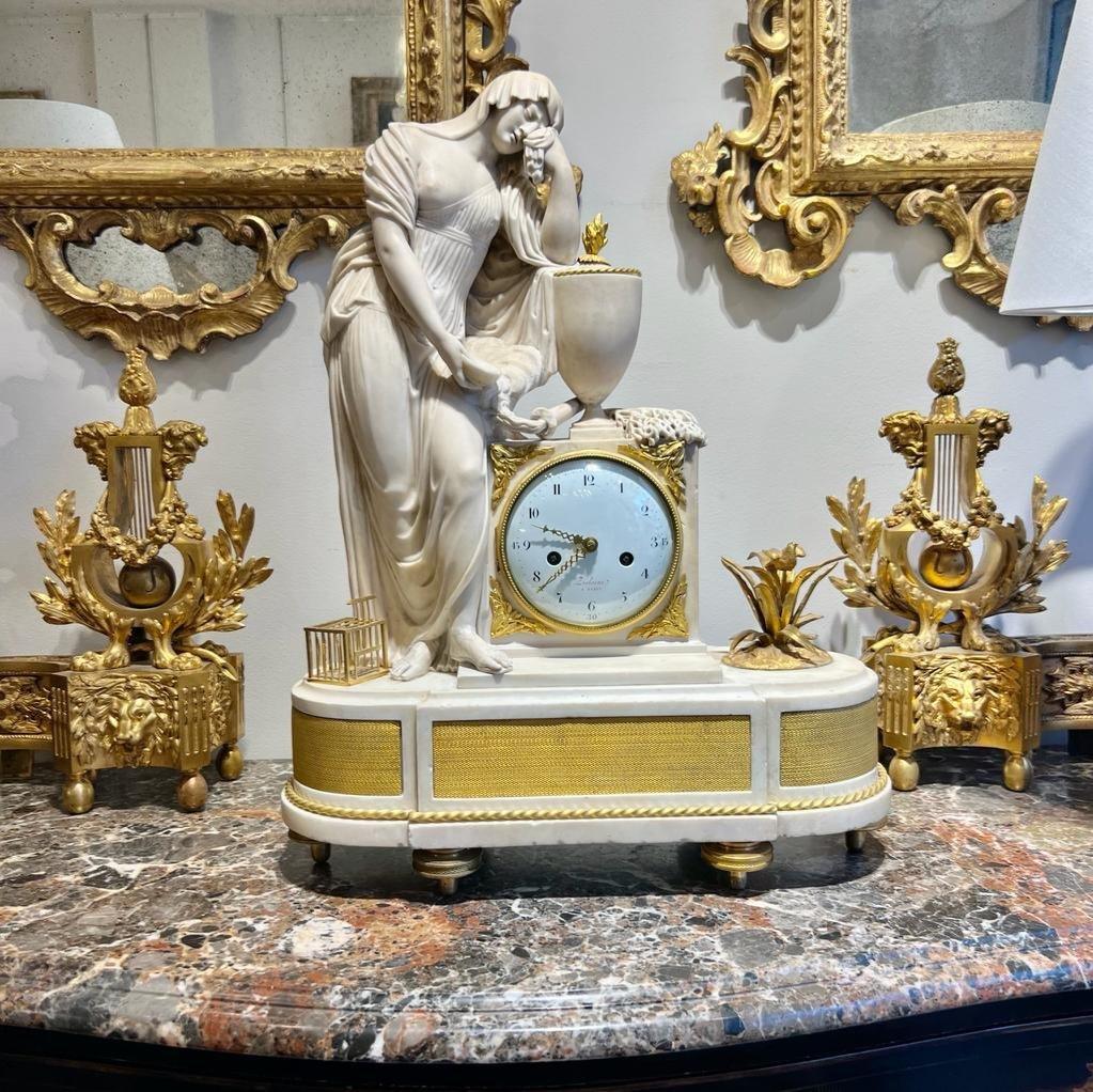 We present you this stunning Carrara marble clock, a masterpiece capturing a statue of a graceful young woman from classical antiquity in exquisite detail. Her foot delicately rests upon the pedestal of the clock, and she is  draped in classical