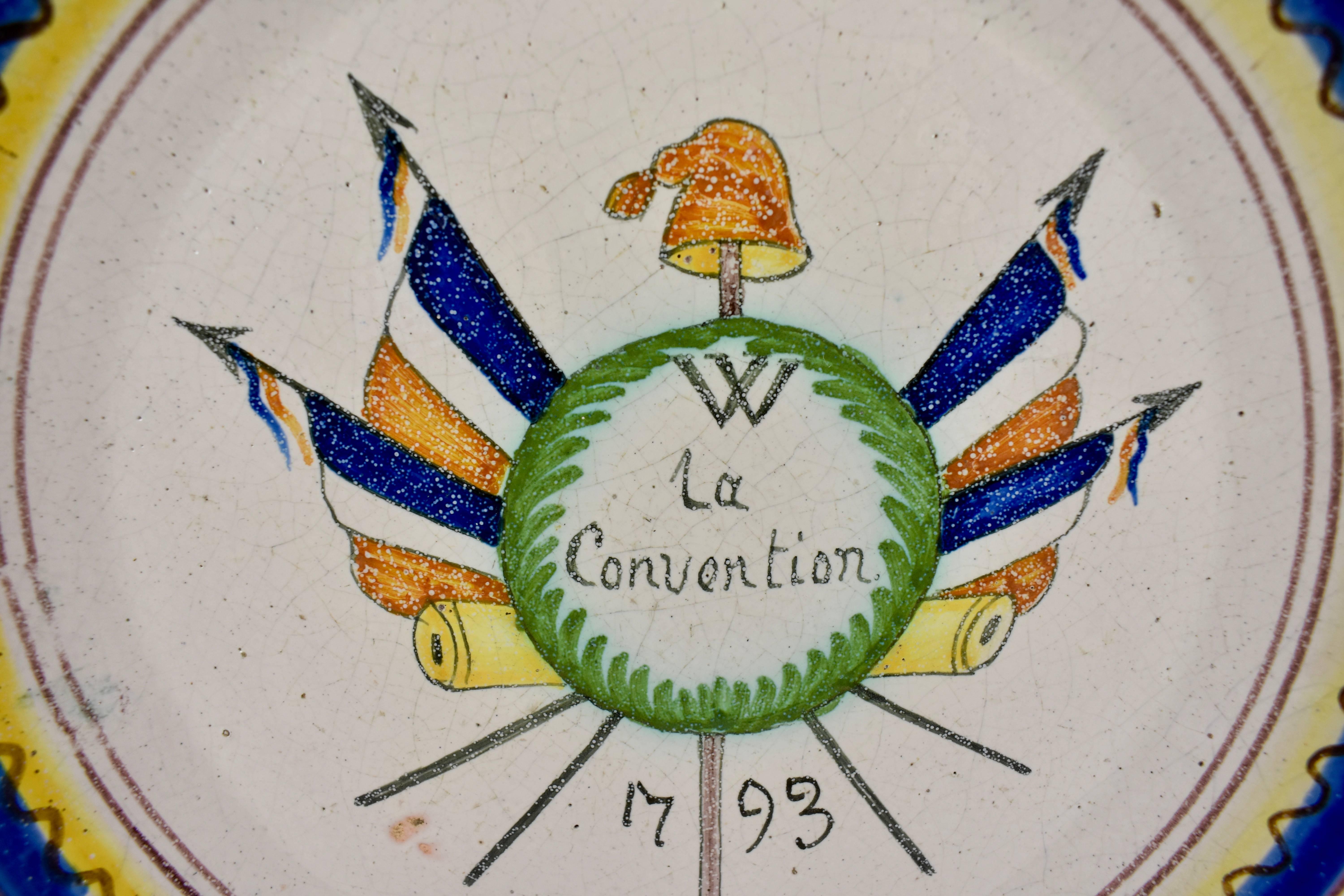 A French Revolution tin-glazed earthenware dish, showing crossed French flags with a Phrygian cap on an épée, the symbol for Liberty. La Convention, meaning The Agreement, is written in script with the date 1793. A hand painted border on the