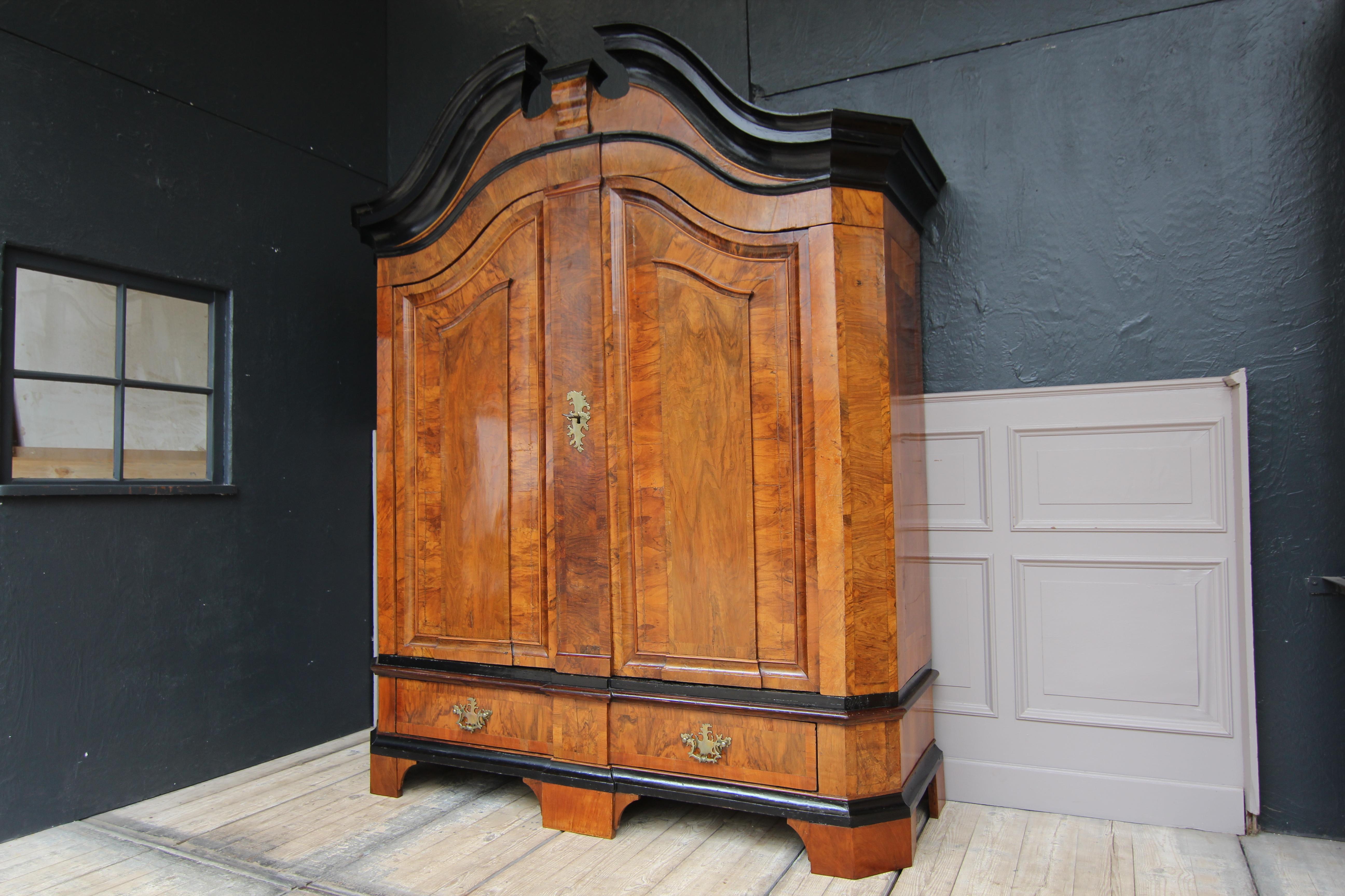 Northern German Baroque cabinet from the late 18th century.
Walnut veneered on oak. Ebonized head strip and profile strips on the base.
Two-door body standing on 5 feet with bevelled corners. Profiled base with large drawer. Curved, blown gable