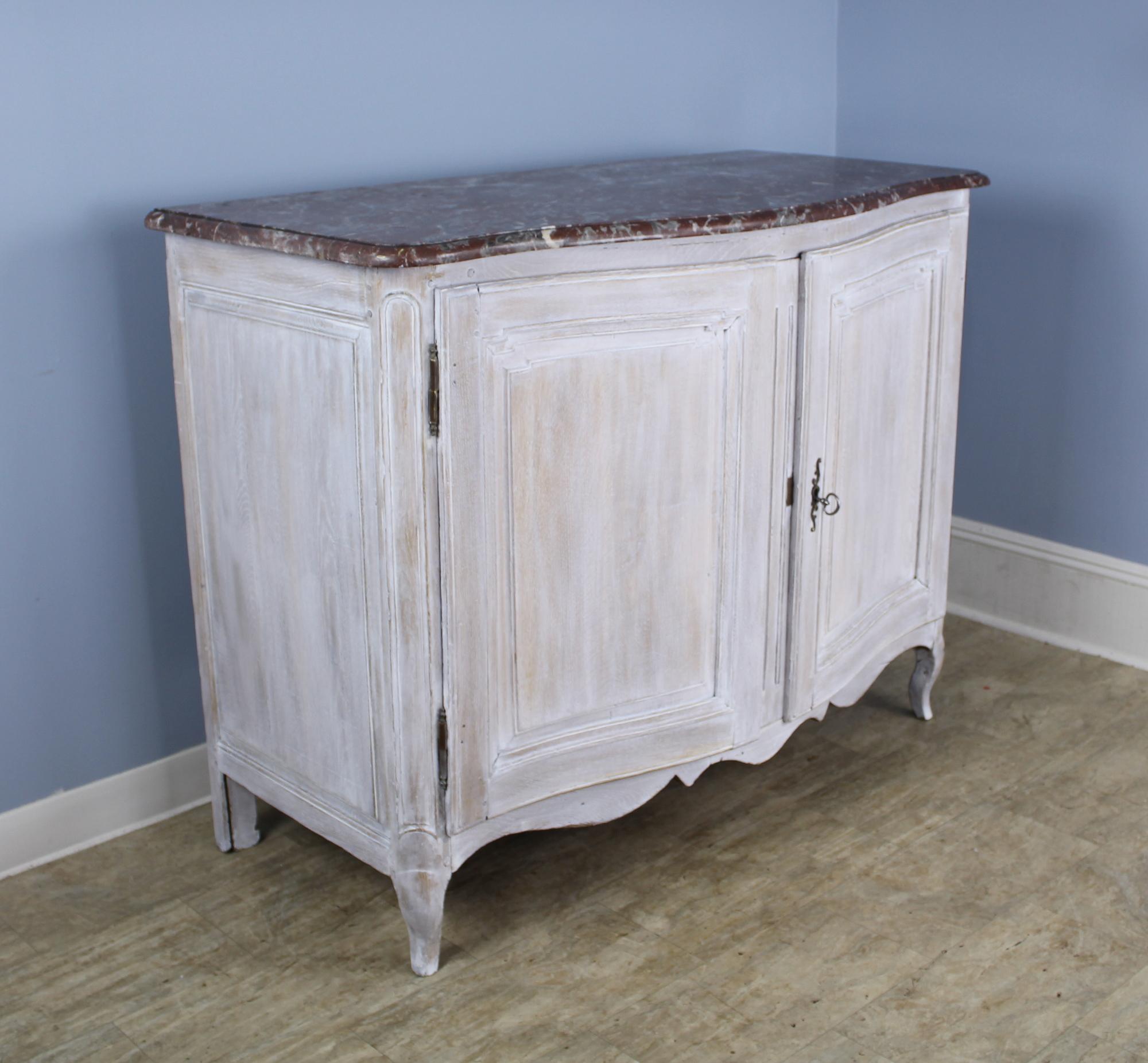 A handsome bowfront buffet, with original dramatically veined marble and newly painted in faux distressed white. Fancifully carved scalloped apron and corner detail. The single interior shelf is not adjustable. The piece closes securely with an