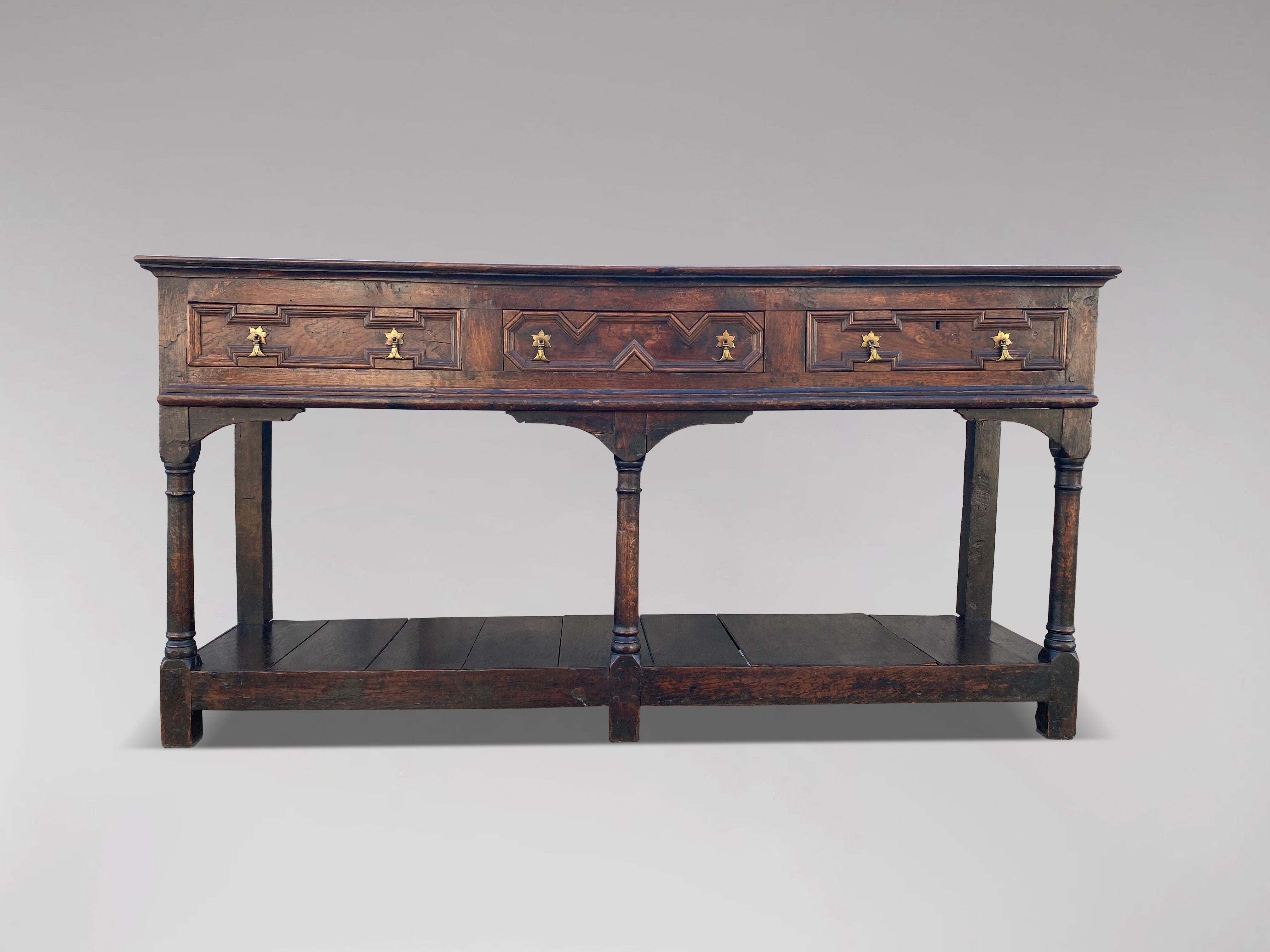 An 18th century oak three drawer open dresser base. Rectangular moulded top above three geometric moulded drawers with small brass drop handles, raised on three turned baluster ring front legs and square back legs and lower pot board shelf. Warm