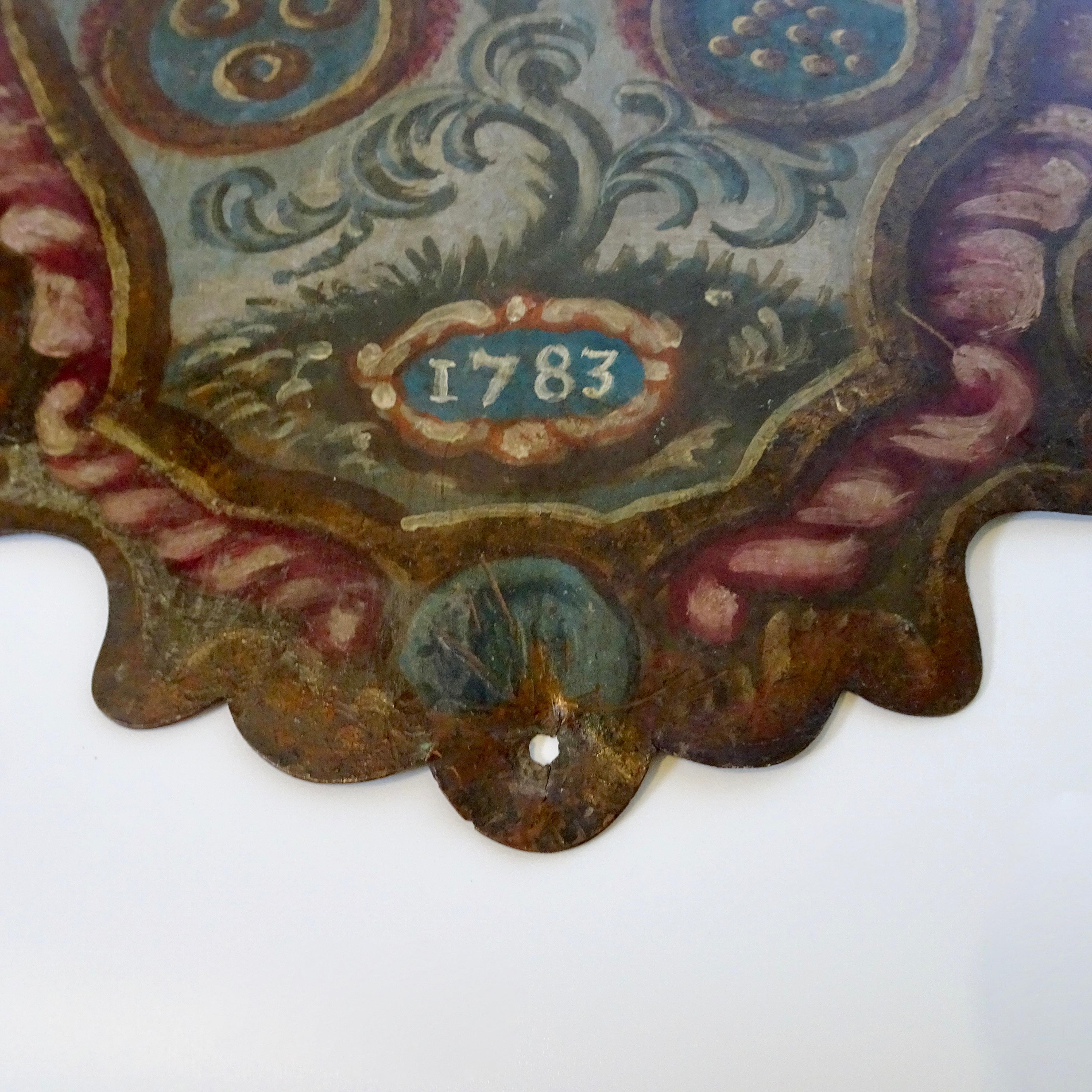 This is a 18th century painted metal shield with a coat of arms. The colors are red, blue and green with a pattern that is similar to a tree of life pattern. The shield has the date 1783 painted.