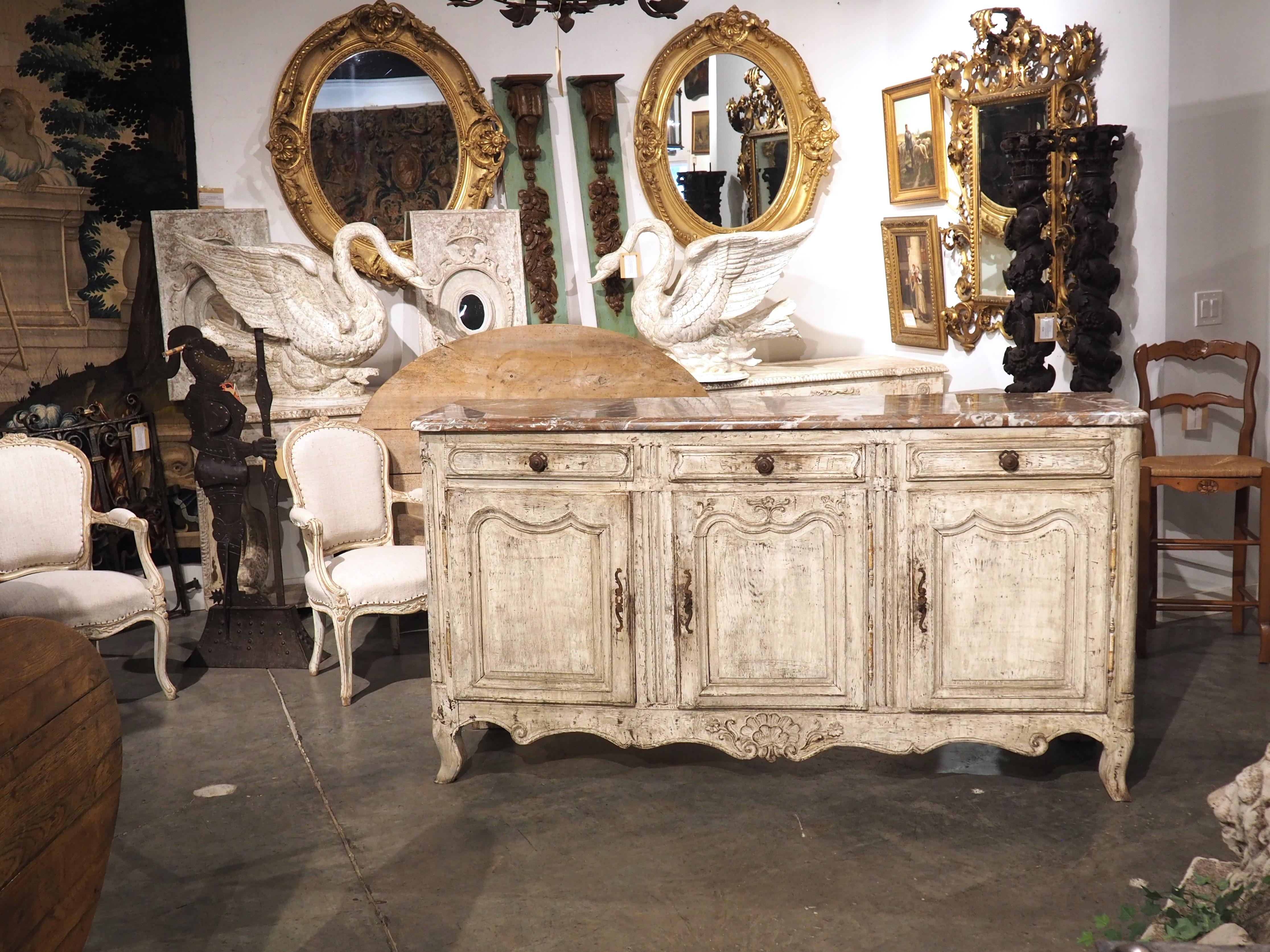 An exquisitely patinated Louis XV enfilade, hand-carved from oak in the 1700s, this large French sideboard features a luxurious 1 1/4″ thick rouge marble top with gray and cream veining, meticulously carved with a quarter-round edge for added