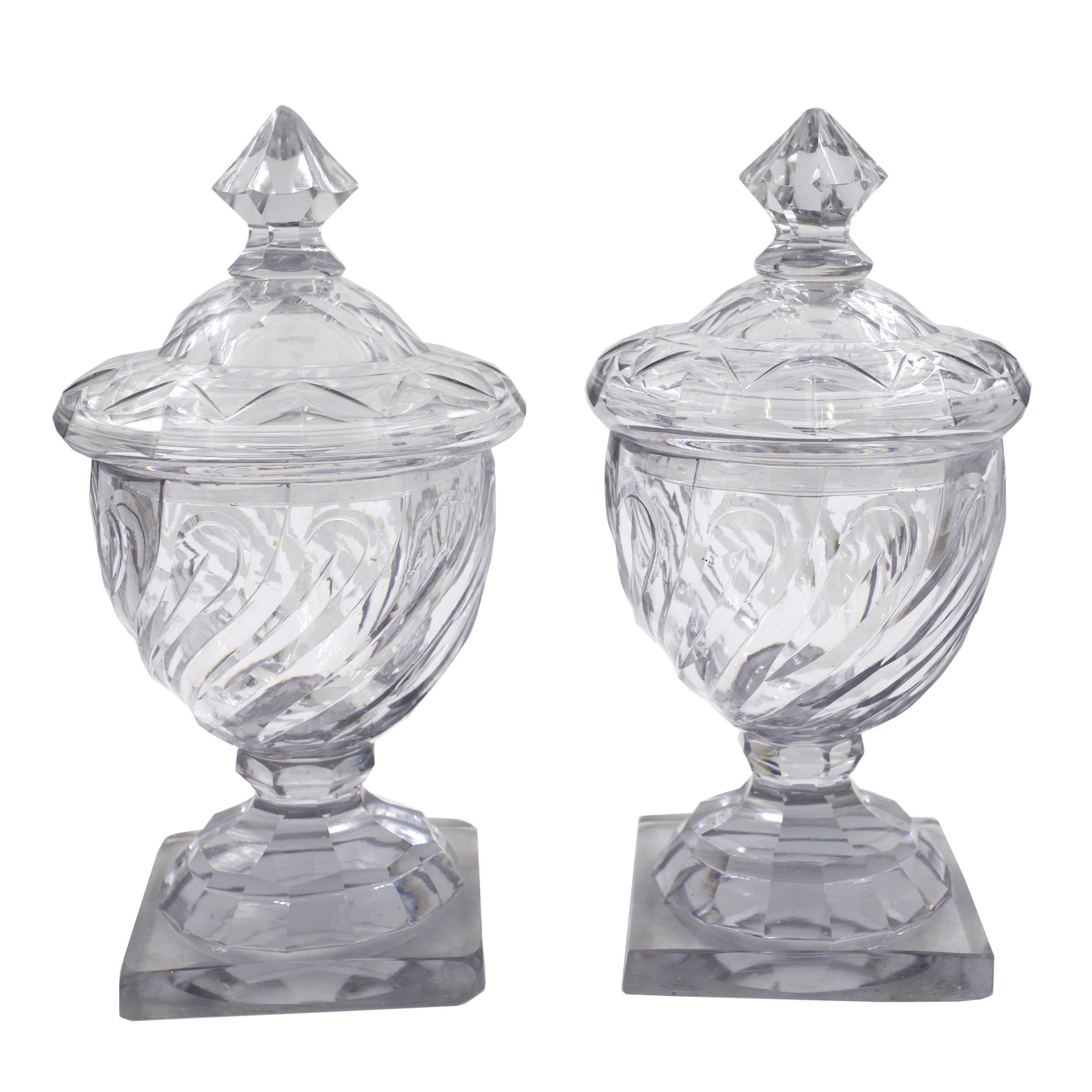 A fine pair of late 18th century Georgian cut glass urns with domed lids, they exhibit a marvelous swirled design in the faceted bodies, the lids with equally geometrical cuts that culminate in cove-facets surrounding the finials. They rest over