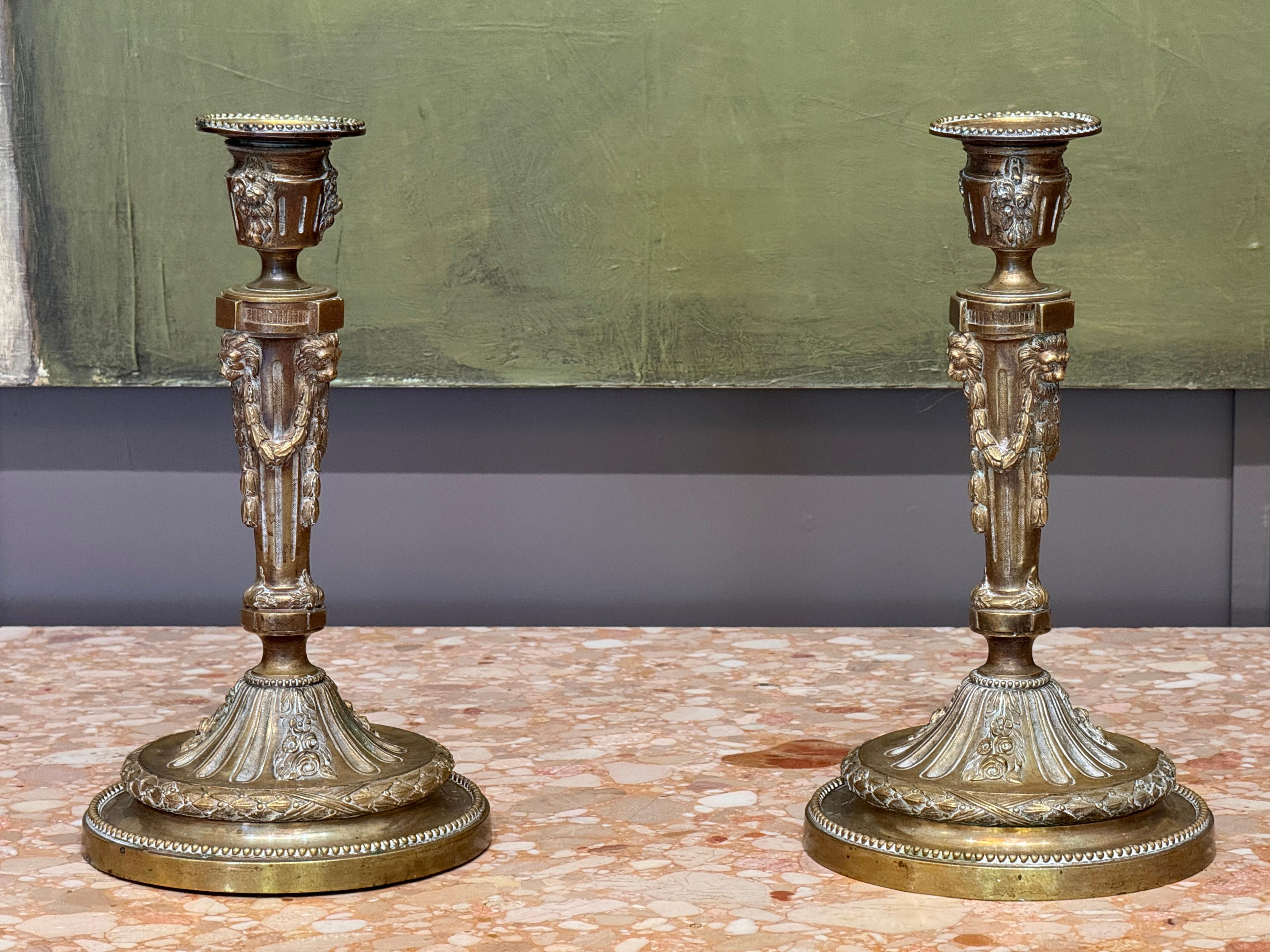 Pair of 18th century French brass candlesticks with classical detailing, featuring lion masks with garland drops at each corner of the faceted stem. The bases are richly ornamented with beaded, floral, and foliate motifs, rising to fluted domed