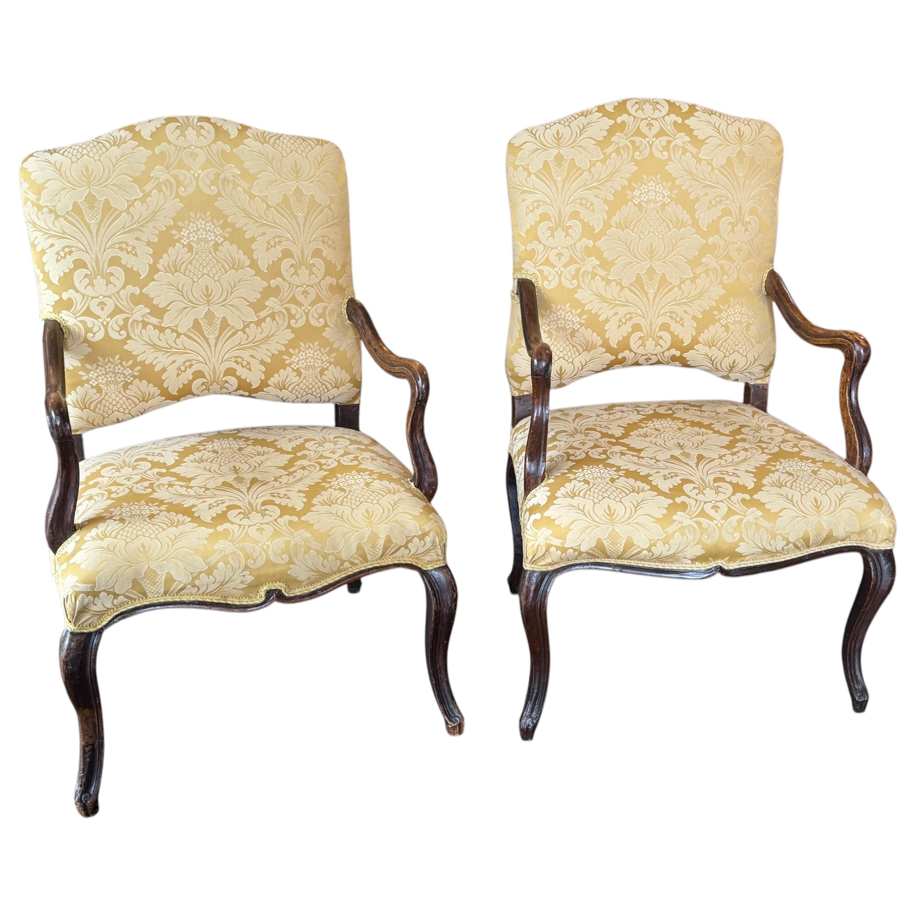 Pair of Louis XV style fauteuils featuring hand-carved walnut frames with shaped arms and cabriole legs. The curved lines and scrollwork reflect the influence of French Rococo design. Upholstered in a patterned gold damask. Elegant and comfortable
